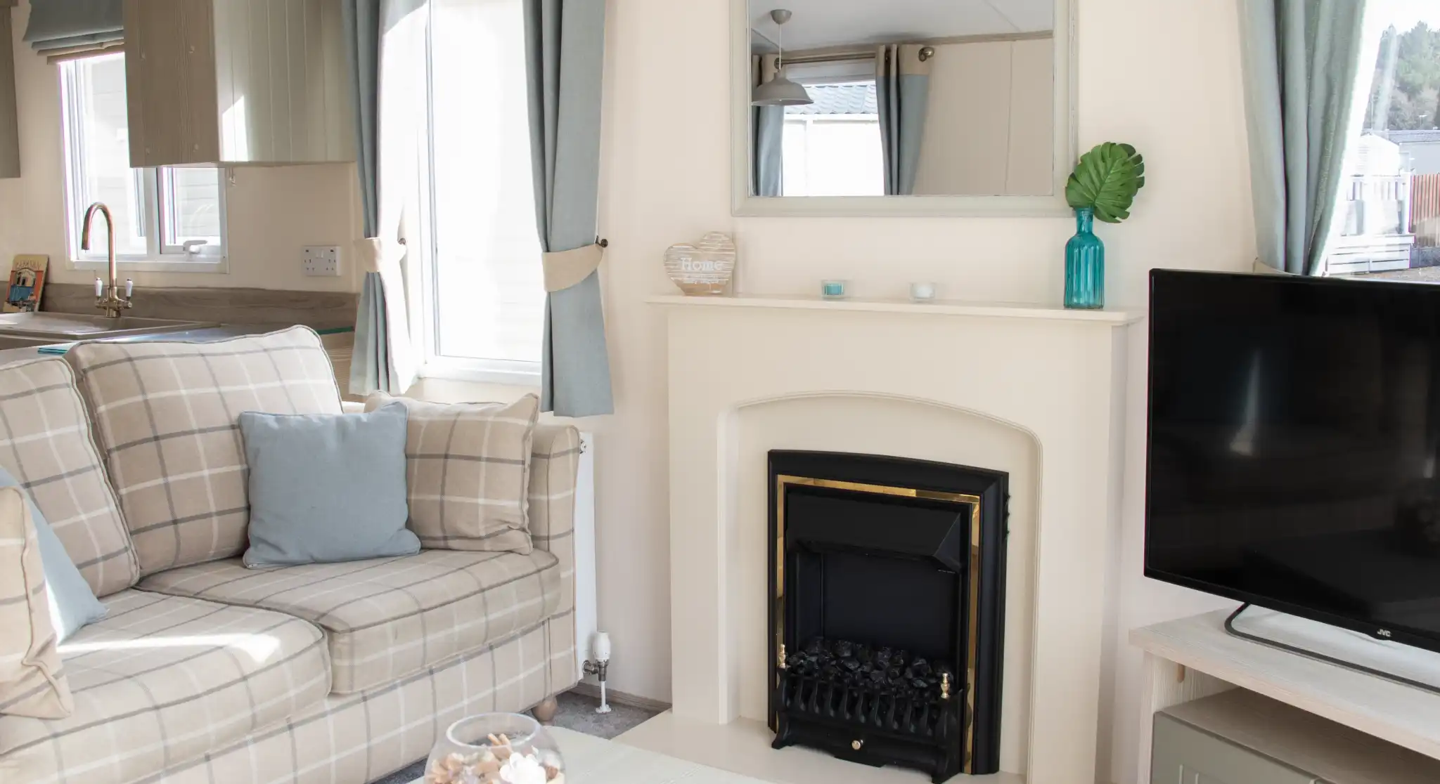 A cozy living room featuring a comfortable plaid sofa with cushions, a small coffee table, and a television. A fireplace is centered on the wall, above which hangs a mirror. Light curtains frame the windows, and decorative items adorn the mantel and table.