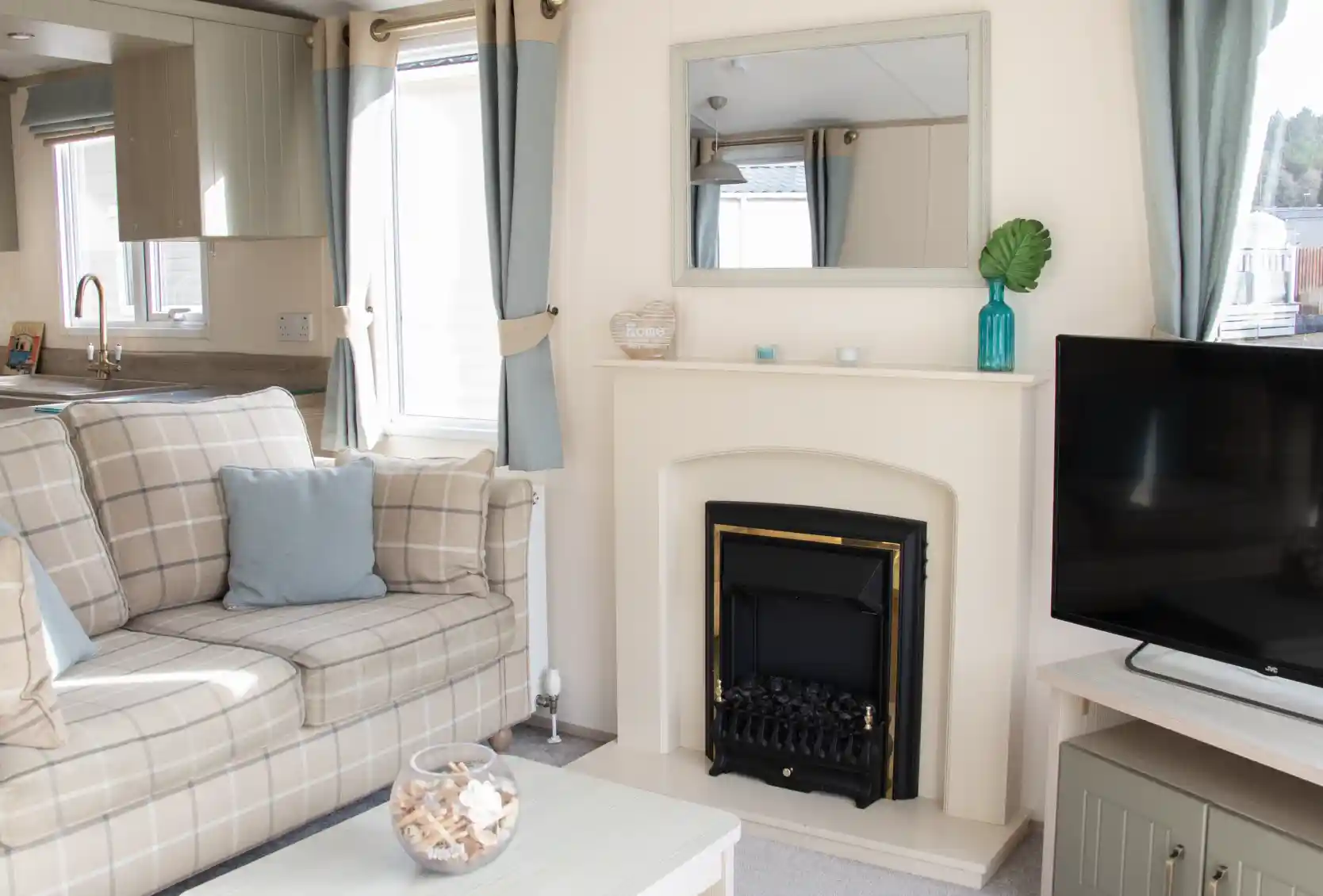 A cozy living room featuring a comfortable plaid sofa with cushions, a small coffee table, and a television. A fireplace is centered on the wall, above which hangs a mirror. Light curtains frame the windows, and decorative items adorn the mantel and table.