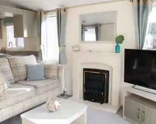 A cozy living room featuring a comfortable plaid sofa with cushions, a small coffee table, and a television. A fireplace is centered on the wall, above which hangs a mirror. Light curtains frame the windows, and decorative items adorn the mantel and table.