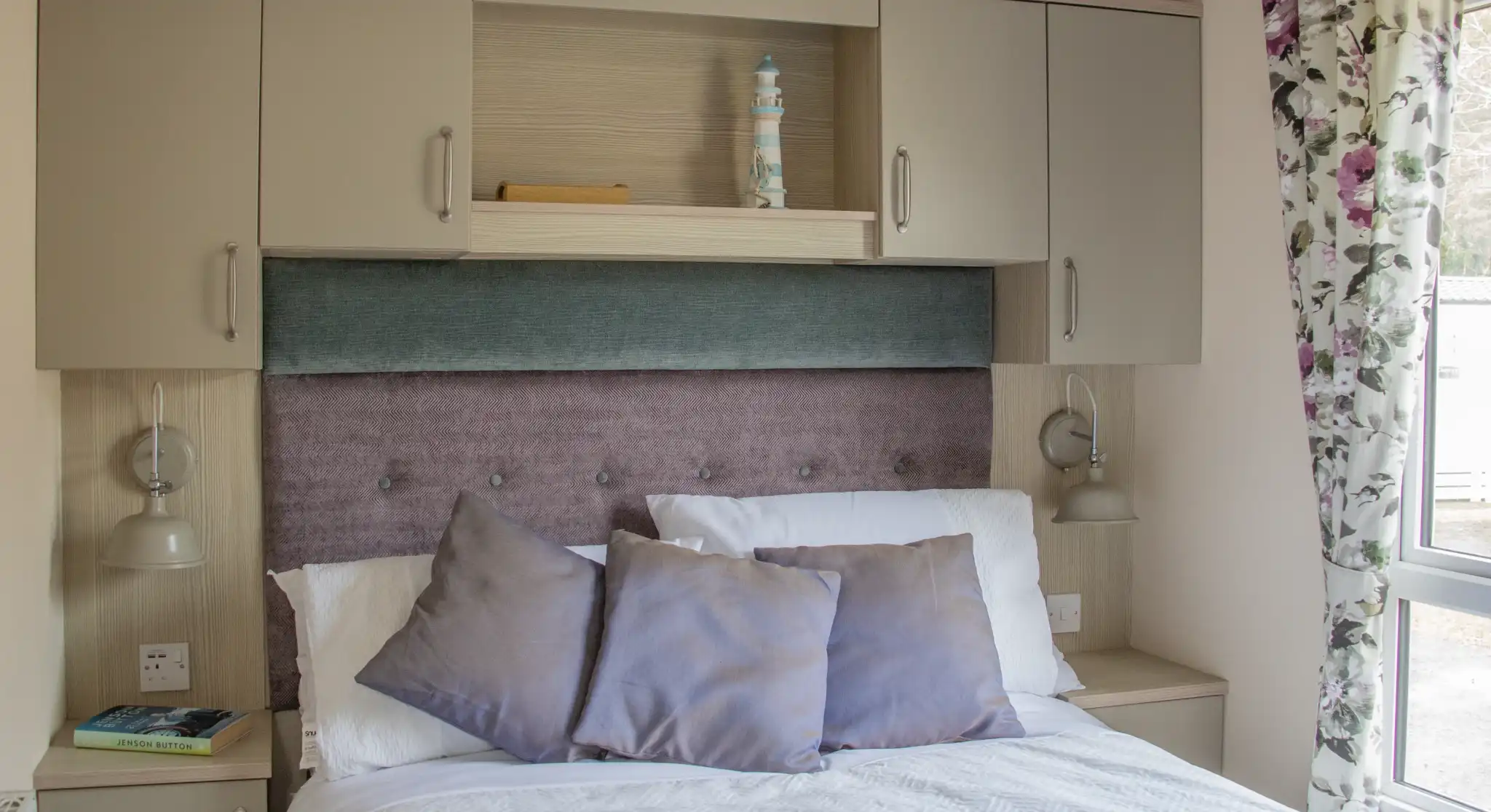 A cozy bedroom featuring a bed with white linens and decorative pillows, a soft purple headboard, and overhead cabinets. Two bedside lamps are mounted next to the bed, and there are floral curtains beside a window, allowing natural light to enter the room.
