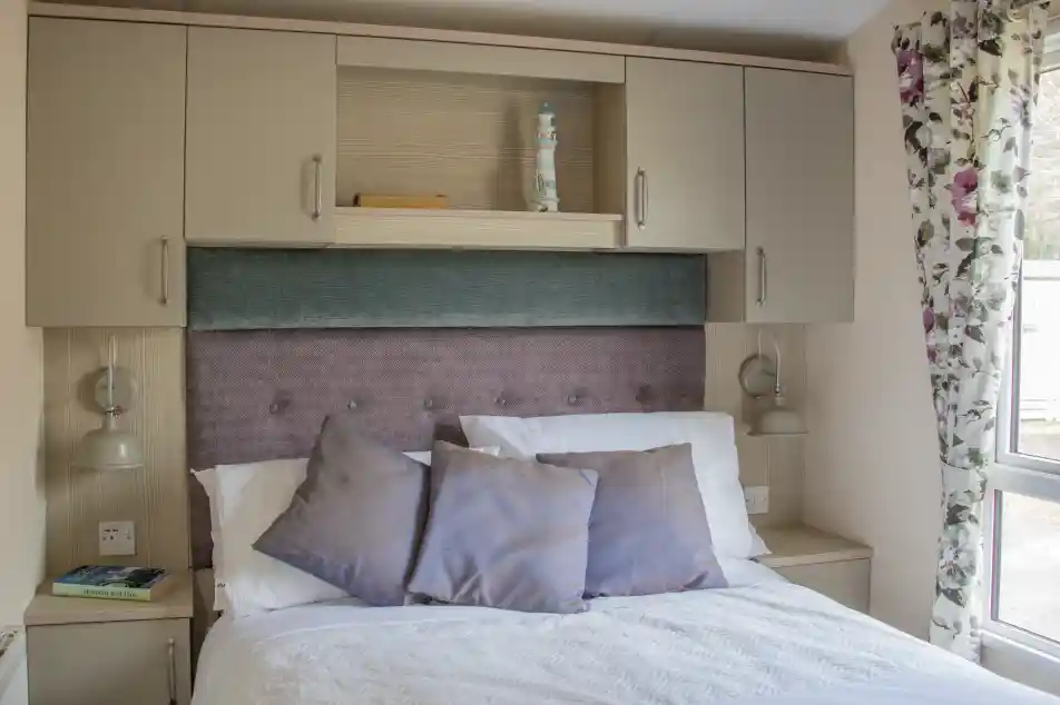 A cozy bedroom featuring a bed with white linens and decorative pillows, a soft purple headboard, and overhead cabinets. Two bedside lamps are mounted next to the bed, and there are floral curtains beside a window, allowing natural light to enter the room.