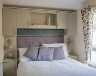 A cozy bedroom featuring a bed with white linens and decorative pillows, a soft purple headboard, and overhead cabinets. Two bedside lamps are mounted next to the bed, and there are floral curtains beside a window, allowing natural light to enter the room.