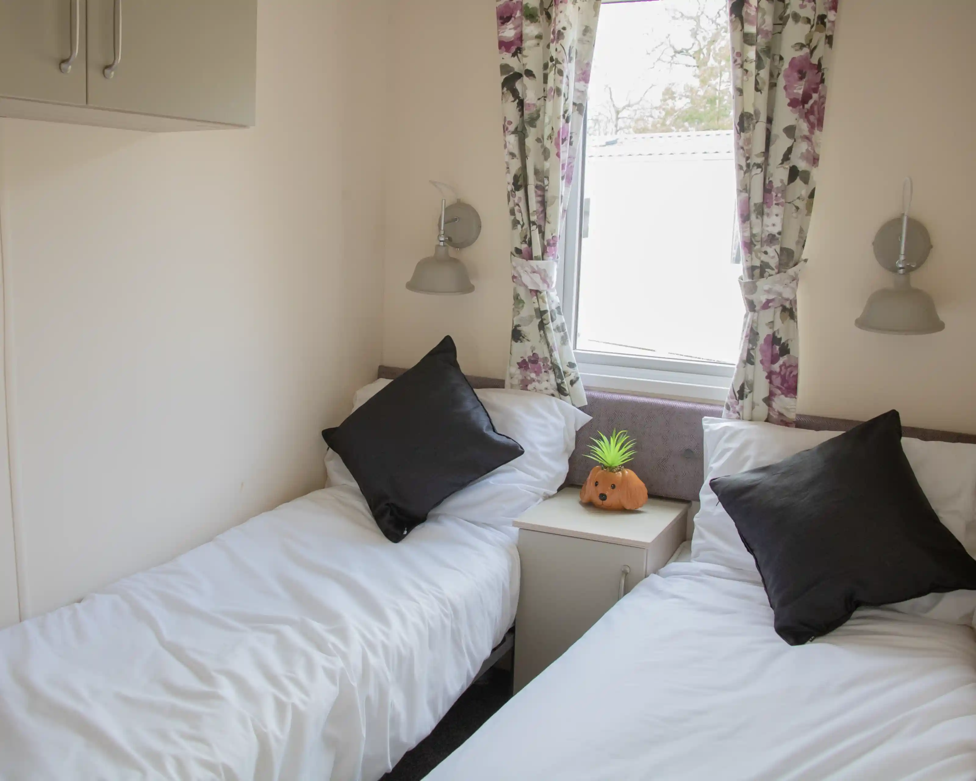 A cozy room with two single beds, each topped with white bedding and black throw pillows. A small nightstand sits between the beds, featuring a decorative orange pumpkin and a green pineapple. Floral curtains frame a window, allowing natural light to illuminate the space.