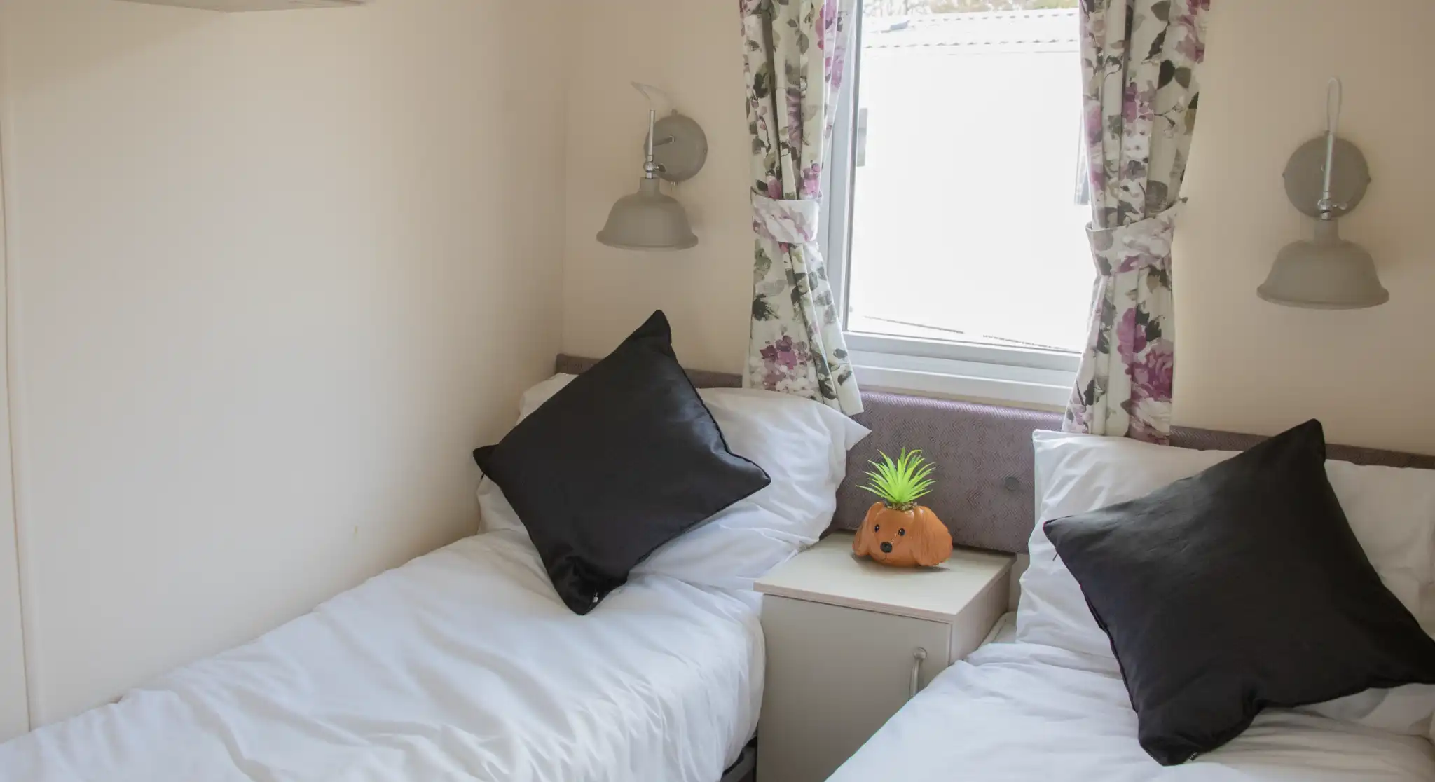 A cozy room with two single beds, each topped with white bedding and black throw pillows. A small nightstand sits between the beds, featuring a decorative orange pumpkin and a green pineapple. Floral curtains frame a window, allowing natural light to illuminate the space.