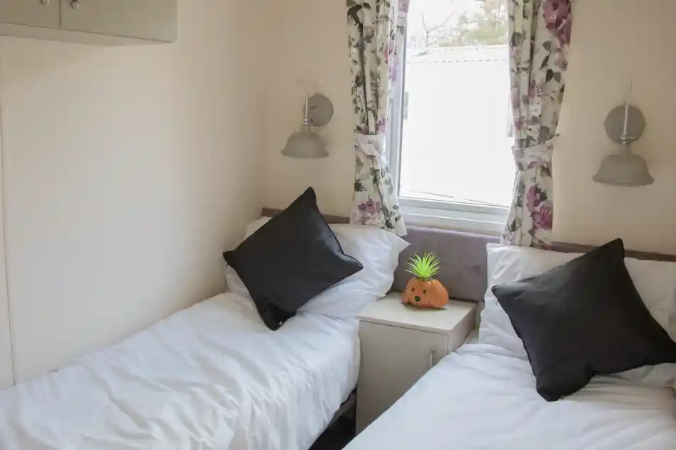 A cozy room with two single beds, each topped with white bedding and black throw pillows. A small nightstand sits between the beds, featuring a decorative orange pumpkin and a green pineapple. Floral curtains frame a window, allowing natural light to illuminate the space.