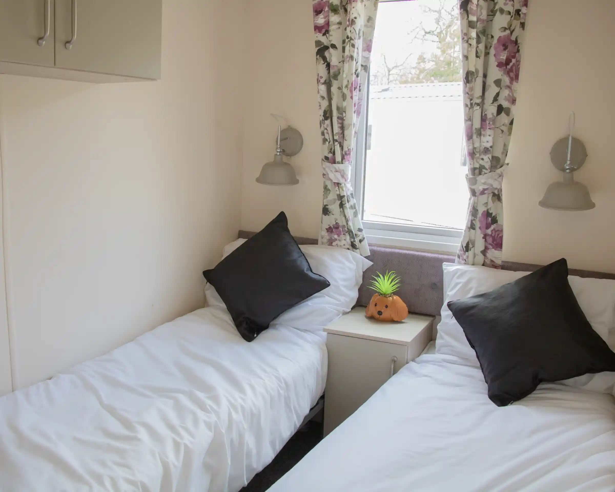 A cozy room with two single beds, each topped with white bedding and black throw pillows. A small nightstand sits between the beds, featuring a decorative orange pumpkin and a green pineapple. Floral curtains frame a window, allowing natural light to illuminate the space.