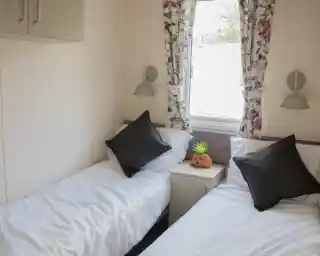 A cozy room with two single beds, each topped with white bedding and black throw pillows. A small nightstand sits between the beds, featuring a decorative orange pumpkin and a green pineapple. Floral curtains frame a window, allowing natural light to illuminate the space.