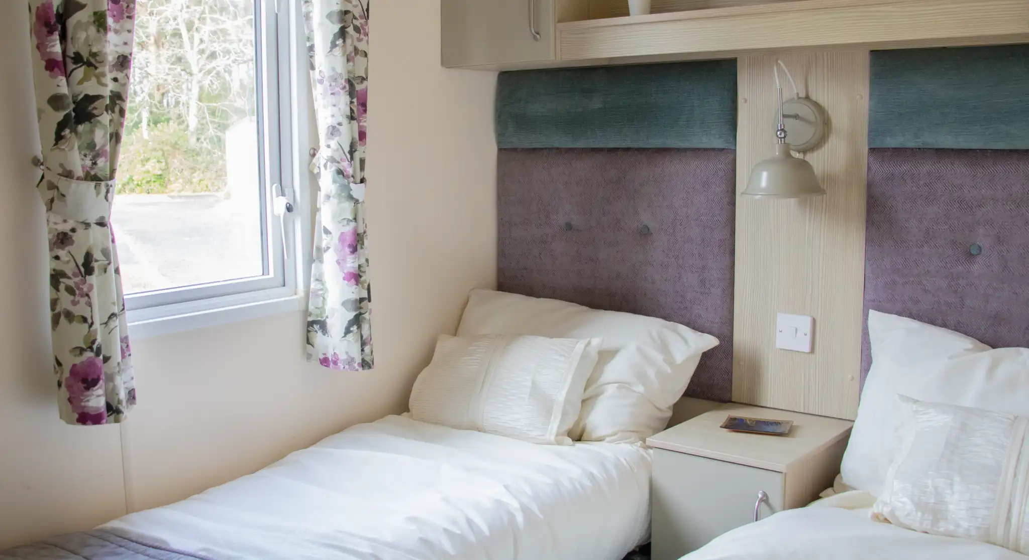 A cozy bedroom featuring two single beds with white linens and gray bedspreads. There's a small nightstand with a plant and two dice on top. A window with floral curtains allows natural light to enter the room.