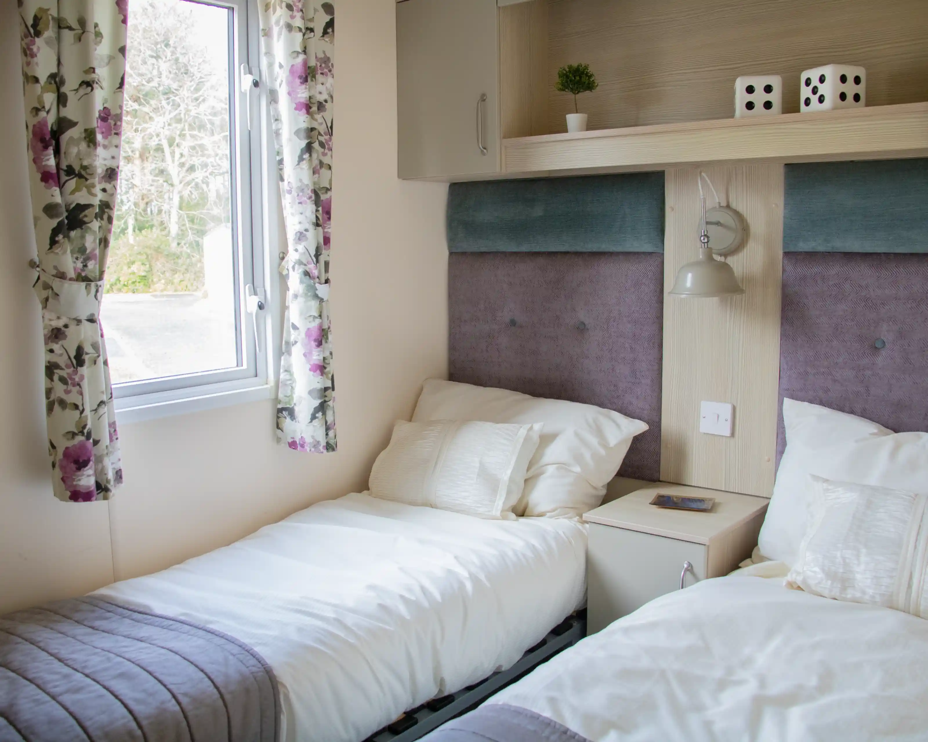 A cozy bedroom featuring two single beds with white linens and gray bedspreads. There's a small nightstand with a plant and two dice on top. A window with floral curtains allows natural light to enter the room.