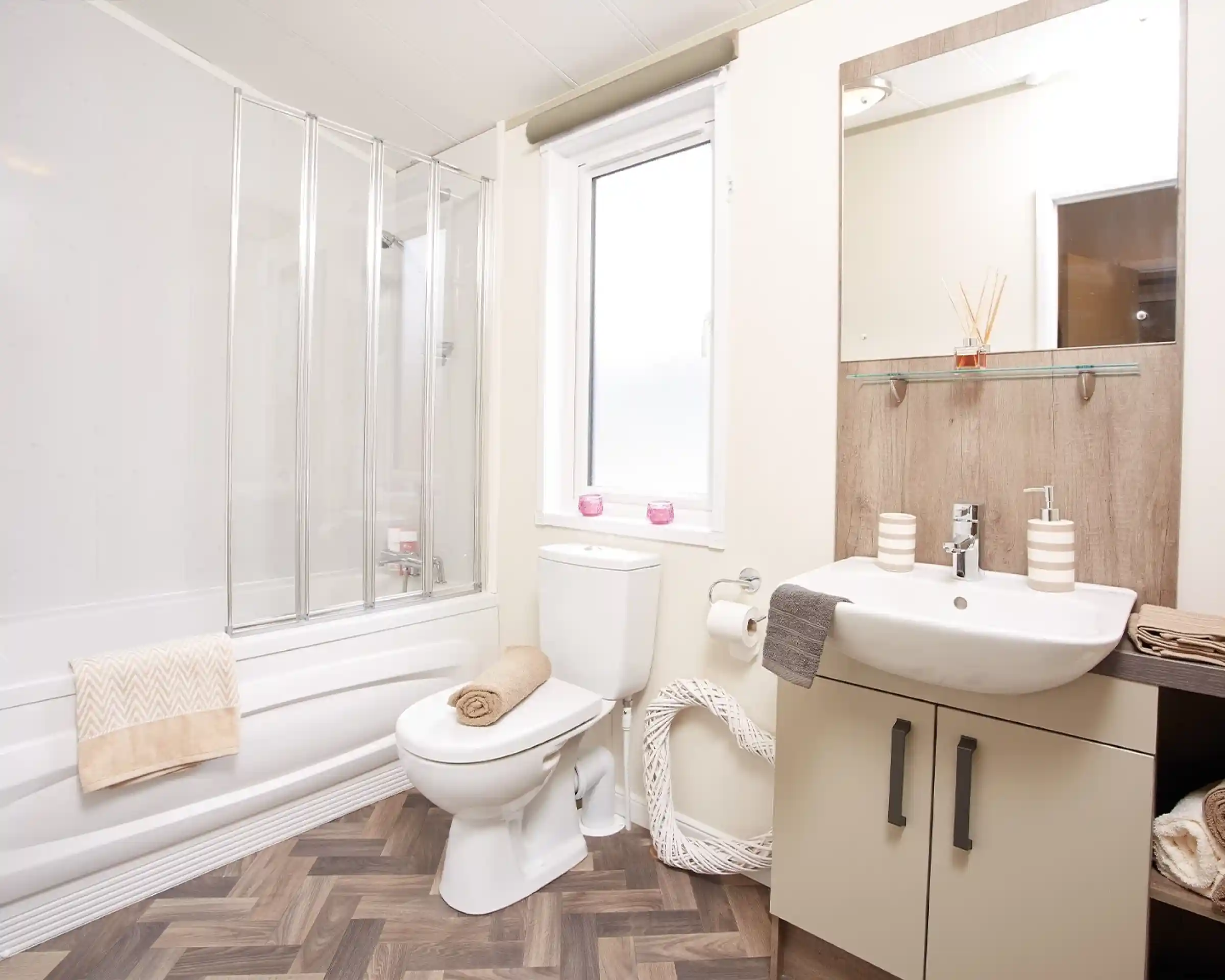 A modern bathroom featuring a shower with glass doors, a white toilet, and a sink with a wooden vanity. Light-colored walls complement the wood-style flooring. Decorative towels are neatly arranged, and a window allows natural light in.