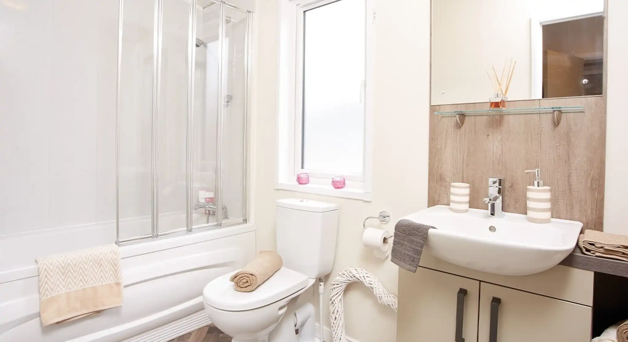 A modern bathroom featuring a shower with glass doors, a white toilet, and a sink with a wooden vanity. Light-colored walls complement the wood-style flooring. Decorative towels are neatly arranged, and a window allows natural light in.