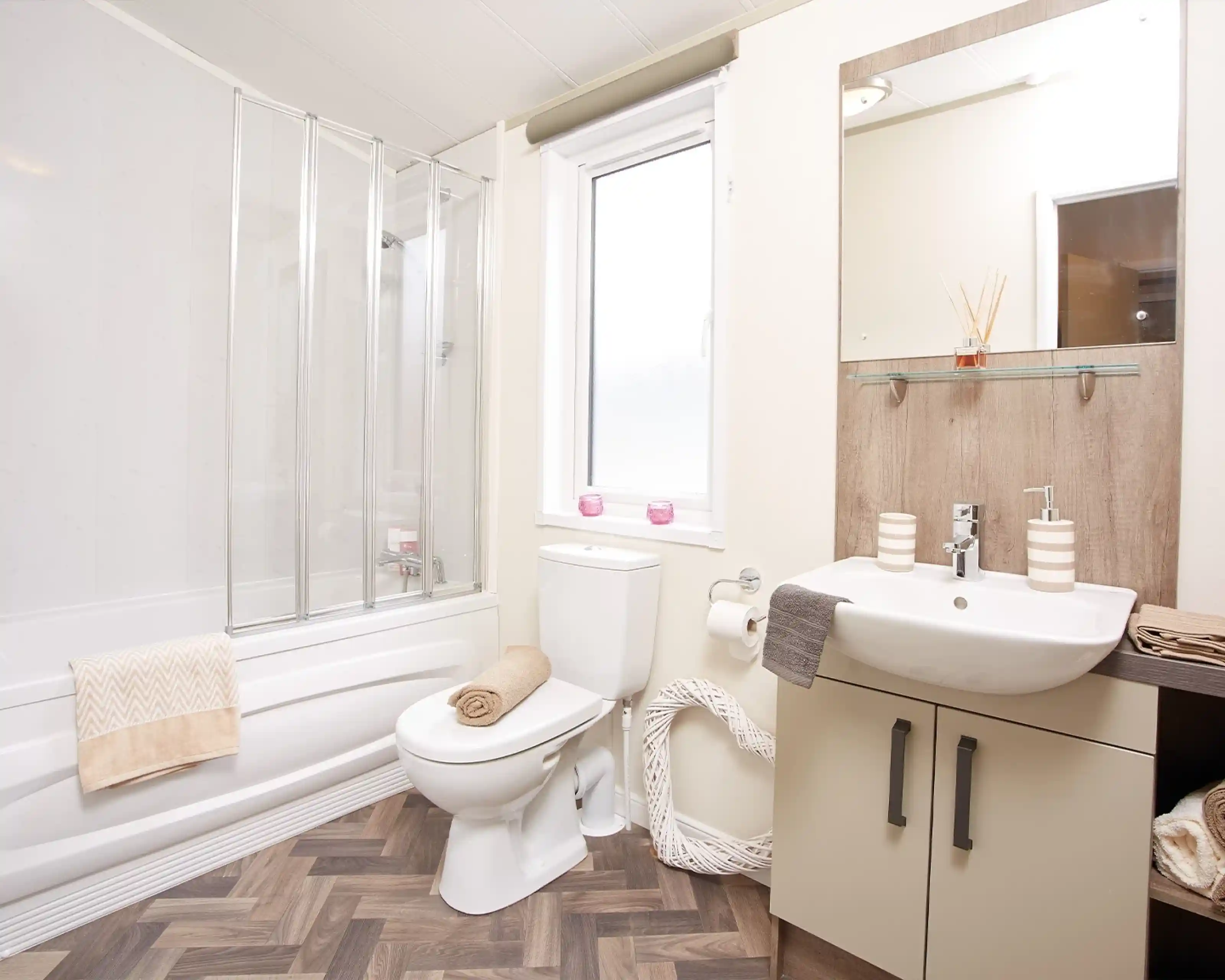A modern bathroom featuring a shower with glass doors, a white toilet, and a sink with a wooden vanity. Light-colored walls complement the wood-style flooring. Decorative towels are neatly arranged, and a window allows natural light in.