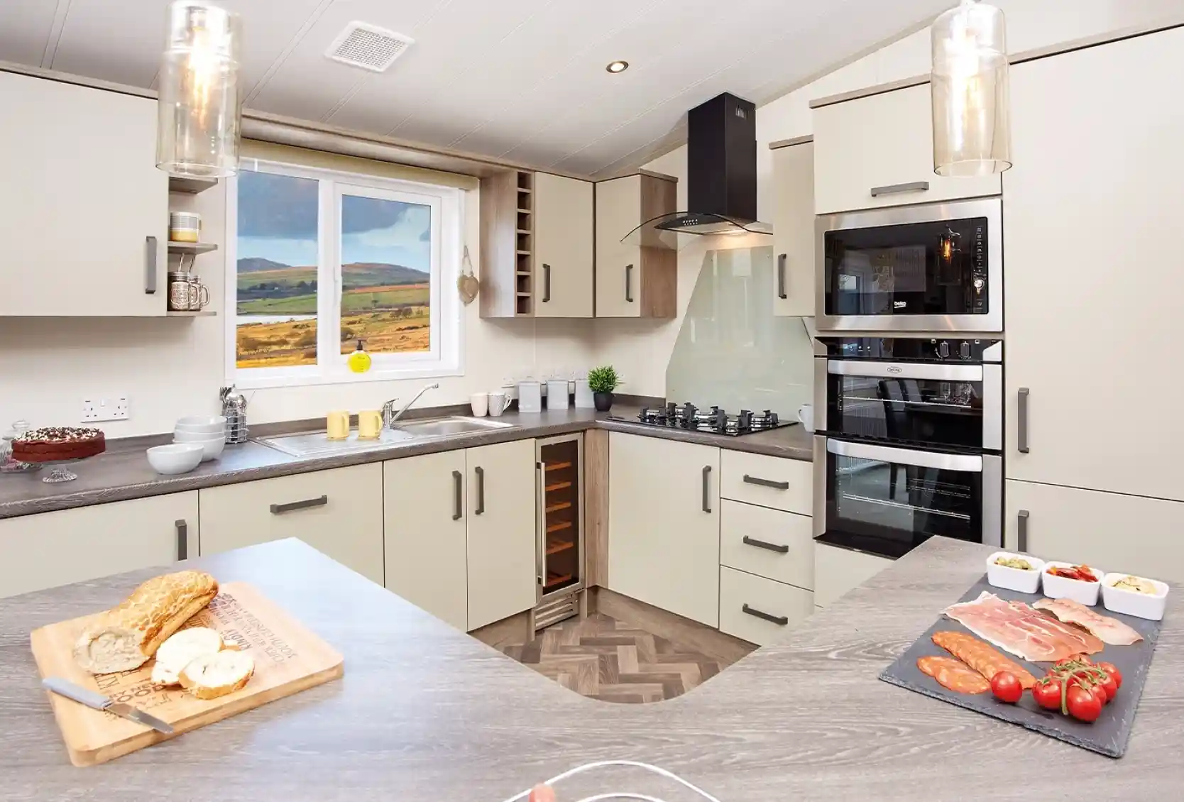Modern kitchen with light-colored cabinetry and a wood-patterned countertop. A window reveals a scenic view of hills. Ingredients for a meal are displayed on the counter, including sliced meats, tomatoes, and a loaf of bread. Stylish pendant lights hang from the ceiling.