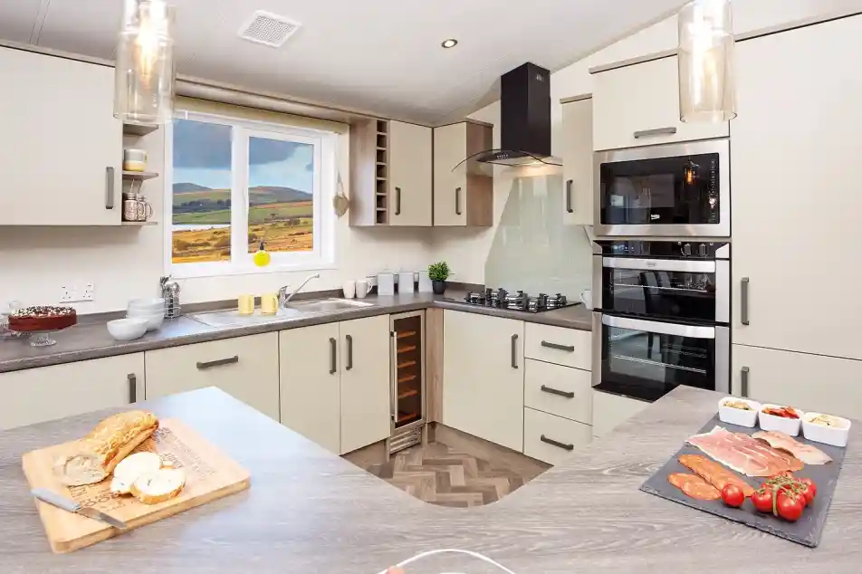 Modern kitchen with light-colored cabinetry and a wood-patterned countertop. A window reveals a scenic view of hills. Ingredients for a meal are displayed on the counter, including sliced meats, tomatoes, and a loaf of bread. Stylish pendant lights hang from the ceiling.