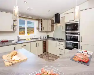 Modern kitchen with light-colored cabinetry and a wood-patterned countertop. A window reveals a scenic view of hills. Ingredients for a meal are displayed on the counter, including sliced meats, tomatoes, and a loaf of bread. Stylish pendant lights hang from the ceiling.