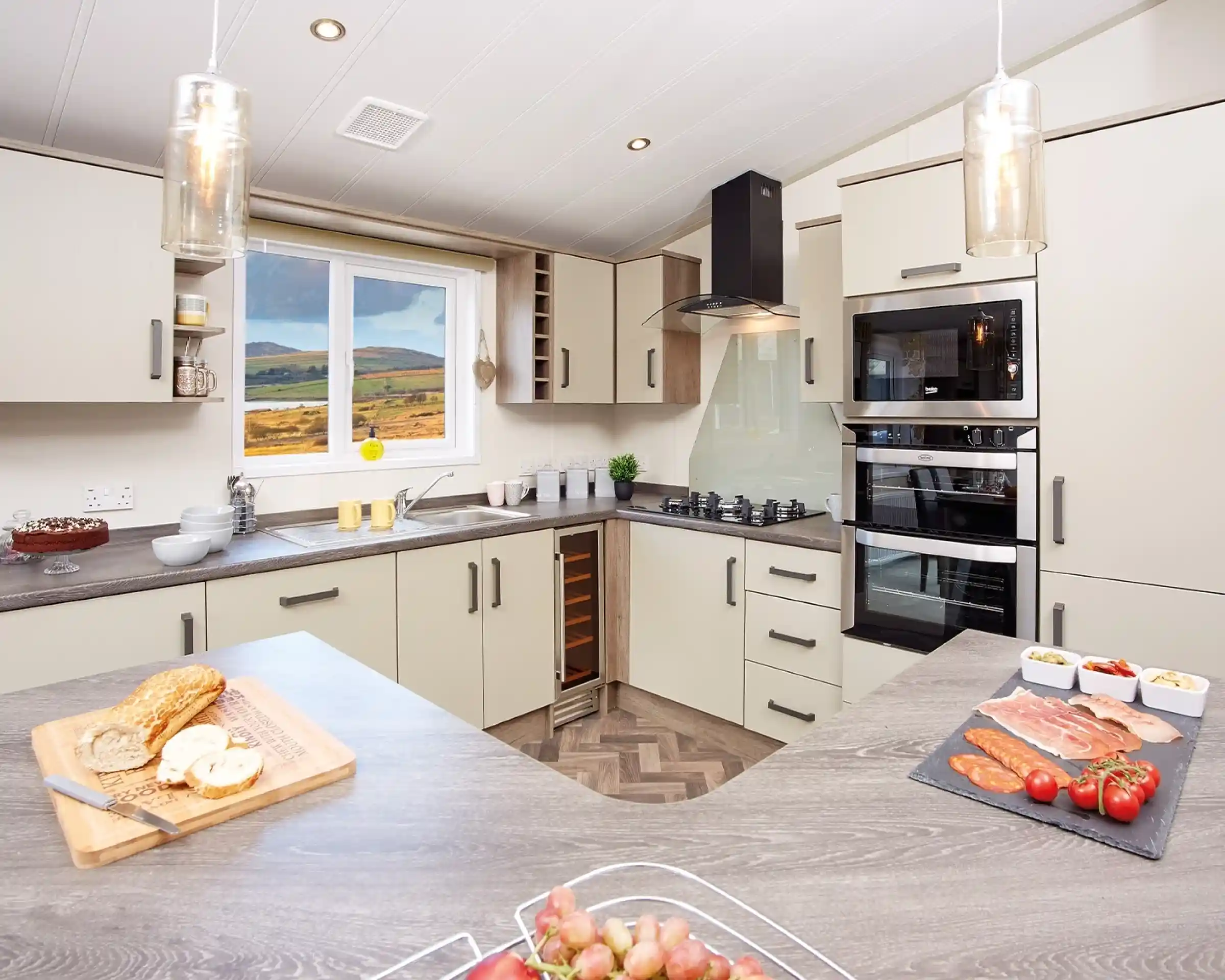 Modern kitchen with light-colored cabinetry and a wood-patterned countertop. A window reveals a scenic view of hills. Ingredients for a meal are displayed on the counter, including sliced meats, tomatoes, and a loaf of bread. Stylish pendant lights hang from the ceiling.
