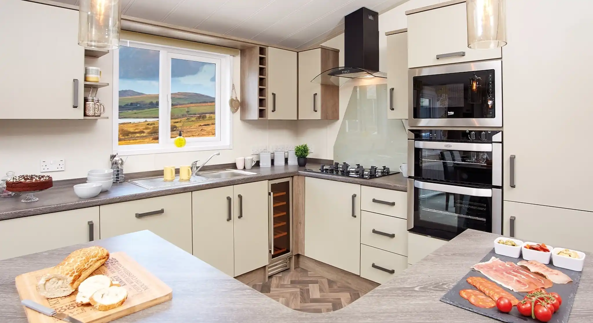 Modern kitchen with light-colored cabinetry and a wood-patterned countertop. A window reveals a scenic view of hills. Ingredients for a meal are displayed on the counter, including sliced meats, tomatoes, and a loaf of bread. Stylish pendant lights hang from the ceiling.