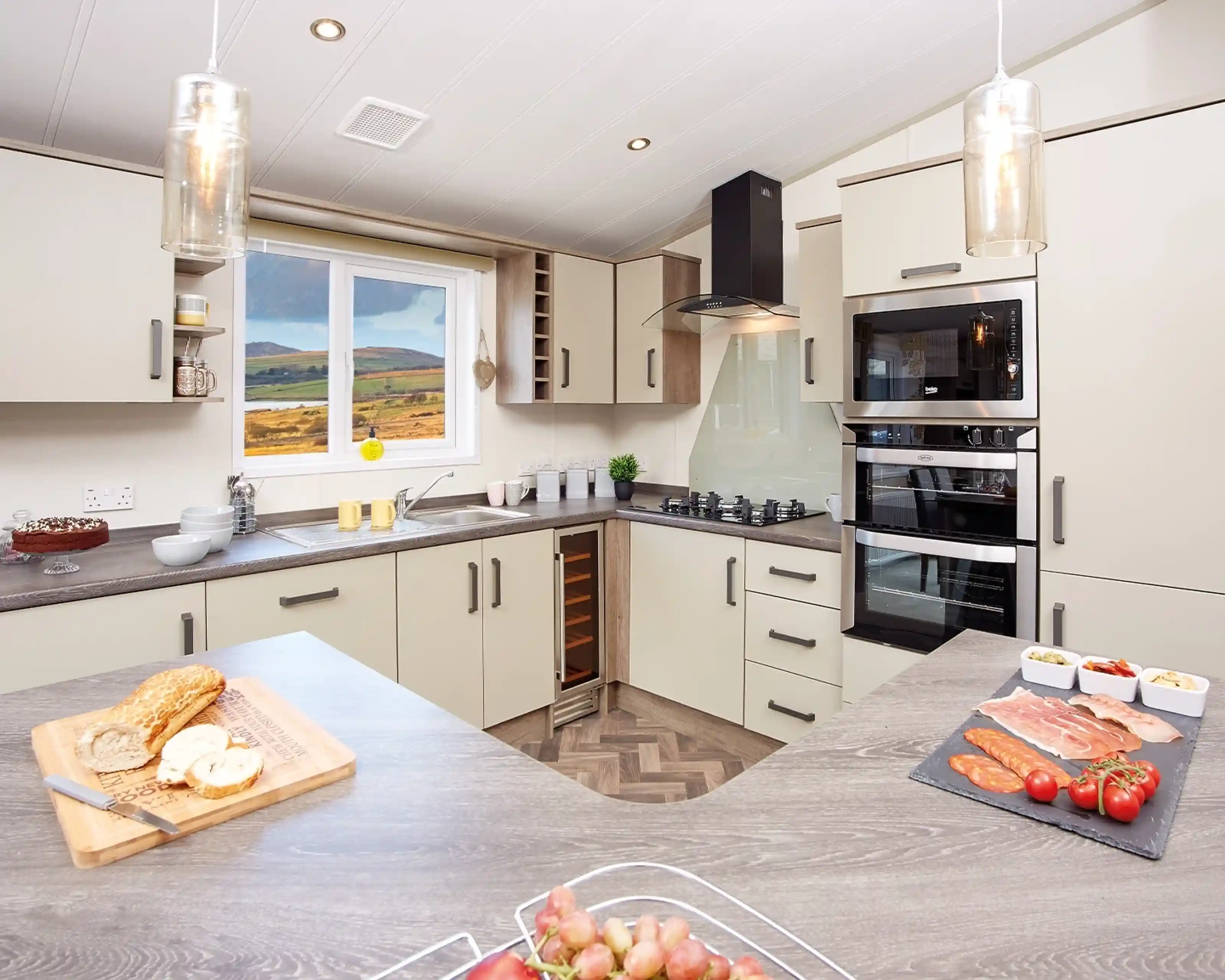 Modern kitchen with light-colored cabinetry and a wood-patterned countertop. A window reveals a scenic view of hills. Ingredients for a meal are displayed on the counter, including sliced meats, tomatoes, and a loaf of bread. Stylish pendant lights hang from the ceiling.