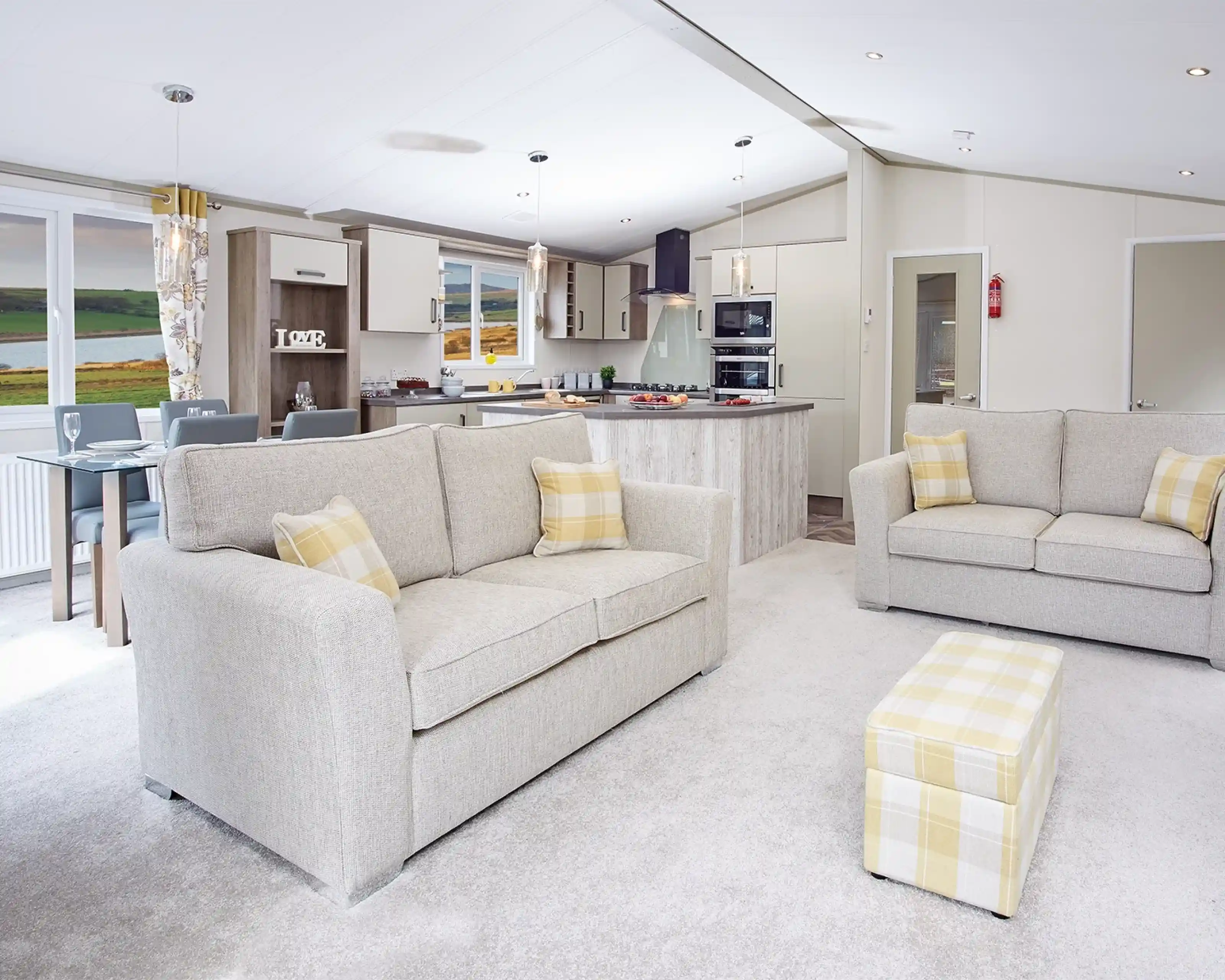 A modern living area featuring two light gray sofas with yellow accent pillows, a small matching ottoman, and a spacious open kitchen in the background. The kitchen includes a large island and contemporary appliances, with a scenic view visible through large windows. Warm, neutral tones dominate the decor.