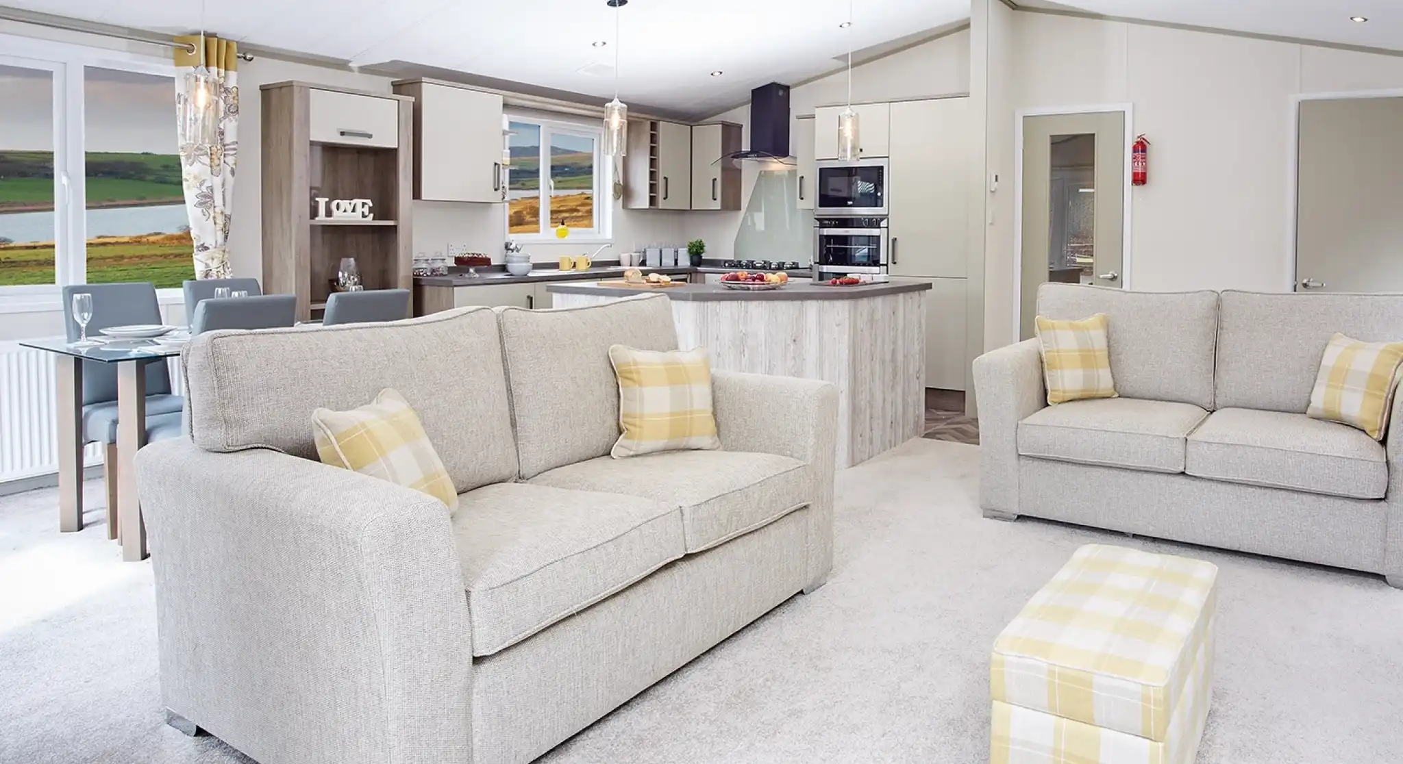 A modern living area featuring two light gray sofas with yellow accent pillows, a small matching ottoman, and a spacious open kitchen in the background. The kitchen includes a large island and contemporary appliances, with a scenic view visible through large windows. Warm, neutral tones dominate the decor.