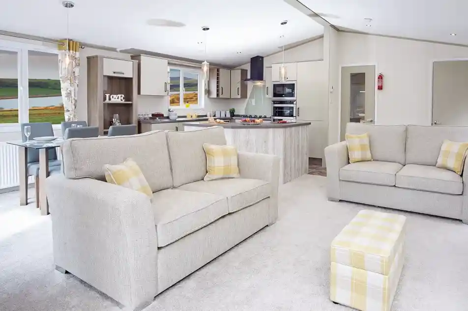 A modern living area featuring two light gray sofas with yellow accent pillows, a small matching ottoman, and a spacious open kitchen in the background. The kitchen includes a large island and contemporary appliances, with a scenic view visible through large windows. Warm, neutral tones dominate the decor.