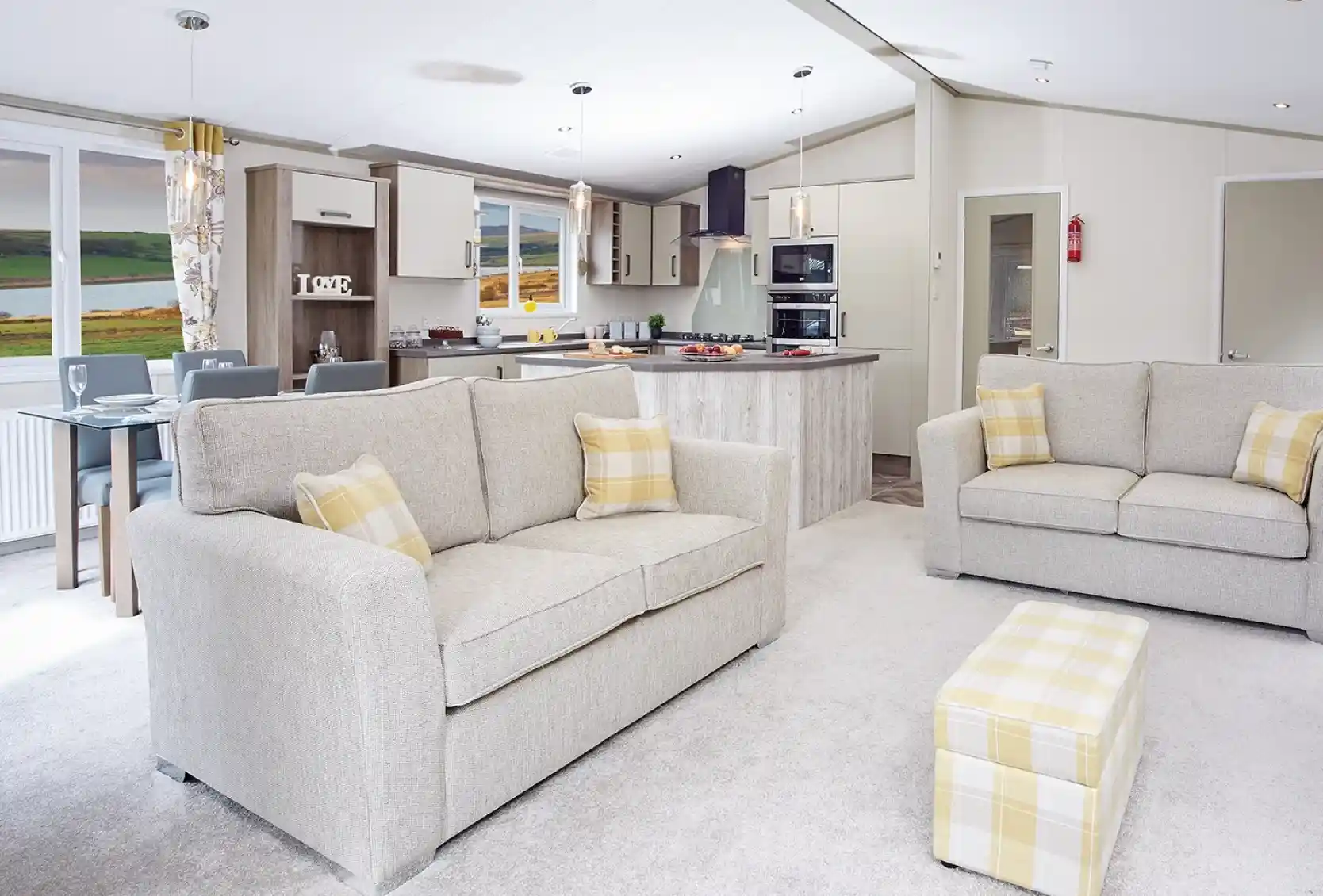 A modern living area featuring two light gray sofas with yellow accent pillows, a small matching ottoman, and a spacious open kitchen in the background. The kitchen includes a large island and contemporary appliances, with a scenic view visible through large windows. Warm, neutral tones dominate the decor.