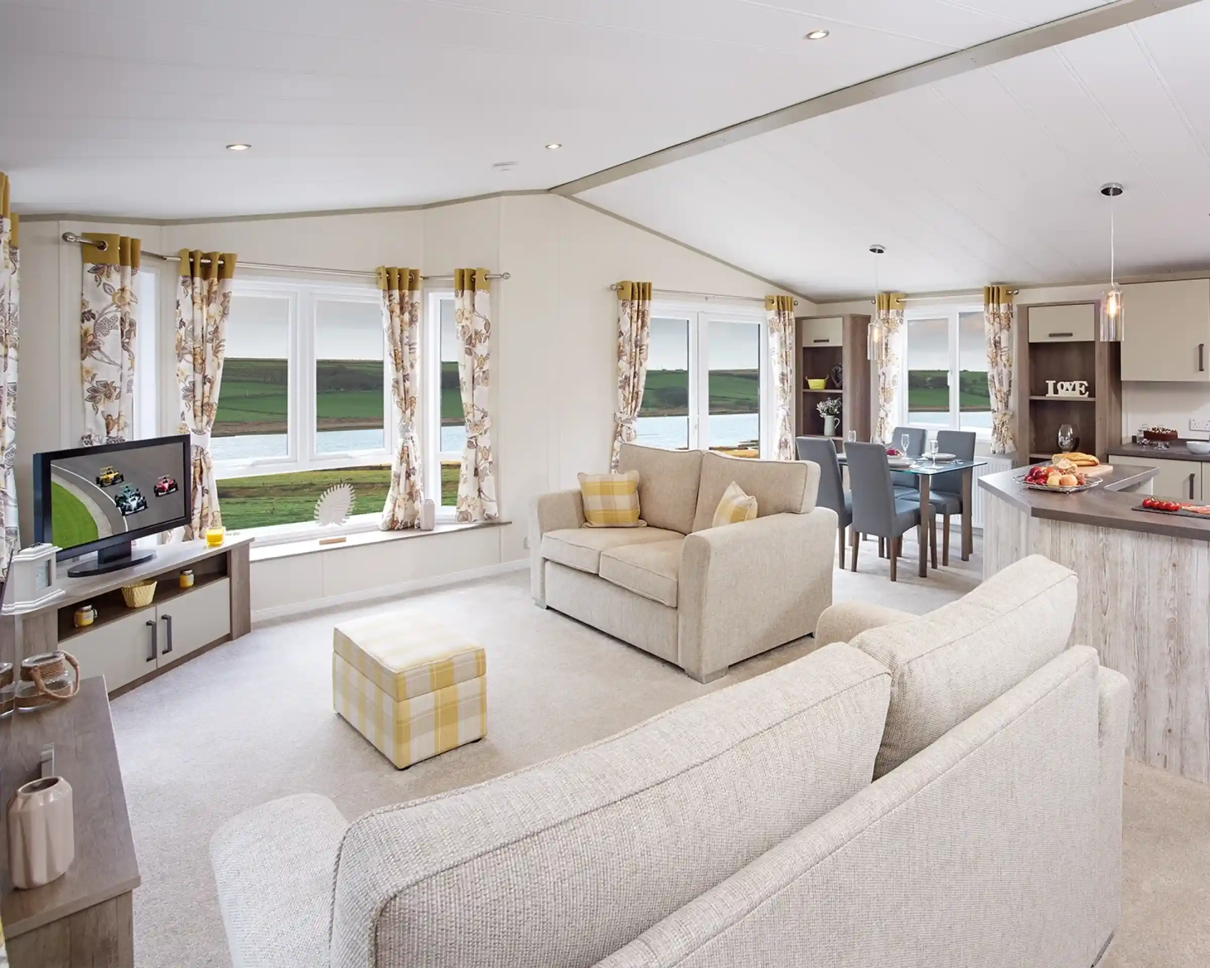A cozy living area featuring a light-colored sofa and an armchair, with large windows offering a scenic view. A TV is positioned on a shelf, and a dining table is set for four. The decor includes floral curtains and soft furnishings. Natural light fills the space, creating a welcoming atmosphere.