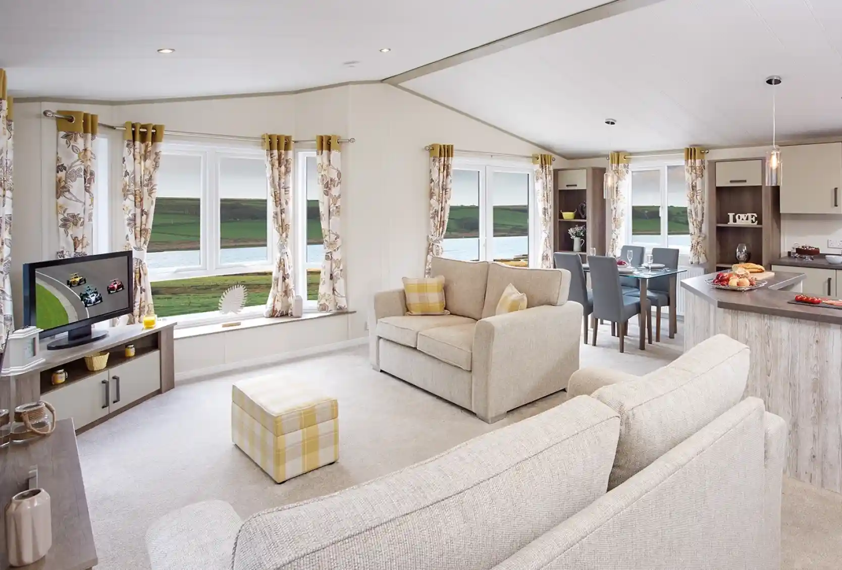 A cozy living area featuring a light-colored sofa and an armchair, with large windows offering a scenic view. A TV is positioned on a shelf, and a dining table is set for four. The decor includes floral curtains and soft furnishings. Natural light fills the space, creating a welcoming atmosphere.