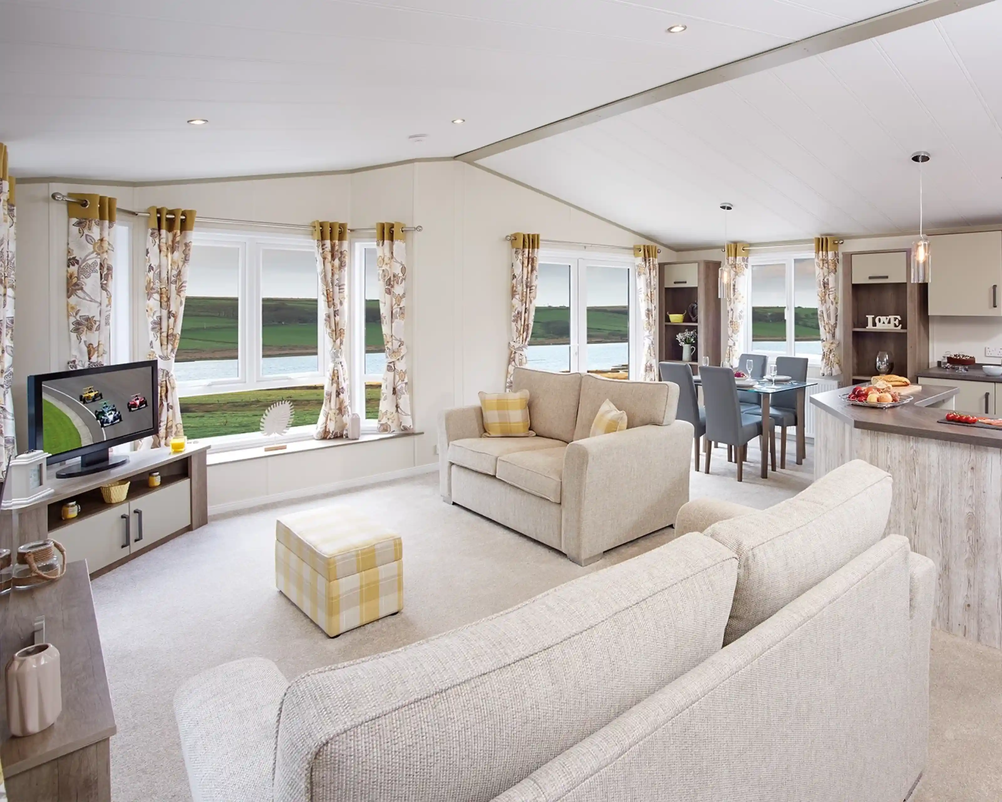 A cozy living area featuring a light-colored sofa and an armchair, with large windows offering a scenic view. A TV is positioned on a shelf, and a dining table is set for four. The decor includes floral curtains and soft furnishings. Natural light fills the space, creating a welcoming atmosphere.