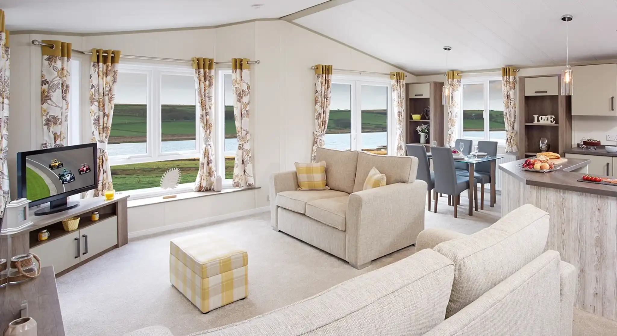 A cozy living area featuring a light-colored sofa and an armchair, with large windows offering a scenic view. A TV is positioned on a shelf, and a dining table is set for four. The decor includes floral curtains and soft furnishings. Natural light fills the space, creating a welcoming atmosphere.