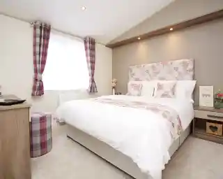 A cozy bedroom featuring a large bed with a floral headboard and white bedding. Two decorative pillows rest on the bed. A small, round stool with a plaid design sits nearby. Natural light filters through a window with sheer curtains, and the walls are softly adorned with light colors and subtle patterns.