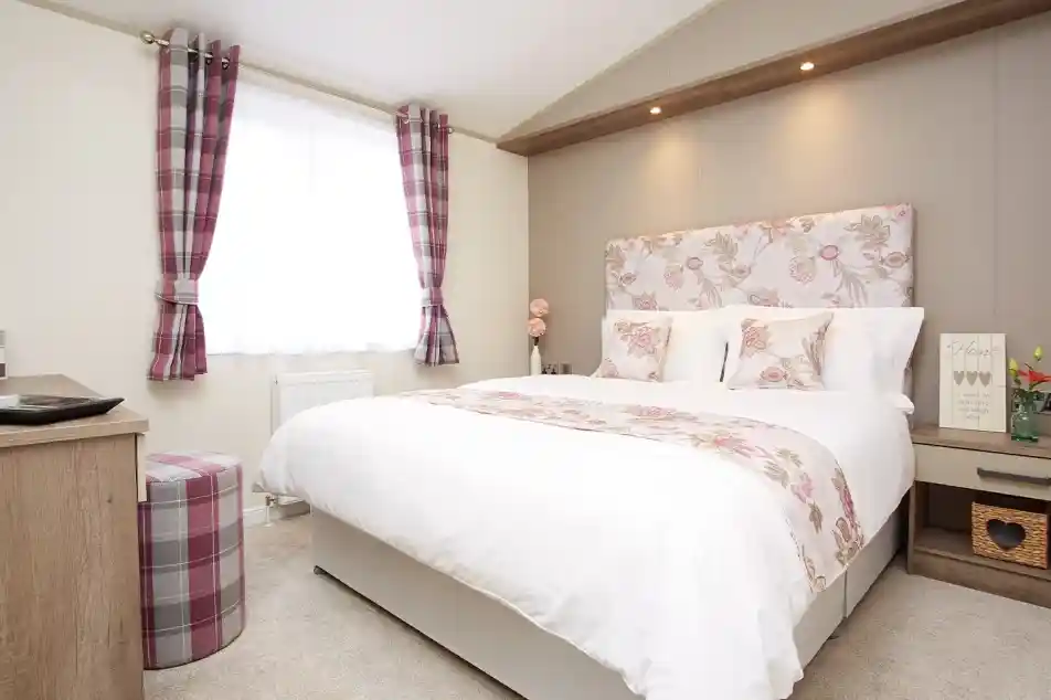 A cozy bedroom featuring a large bed with a floral headboard and white bedding. Two decorative pillows rest on the bed. A small, round stool with a plaid design sits nearby. Natural light filters through a window with sheer curtains, and the walls are softly adorned with light colors and subtle patterns.