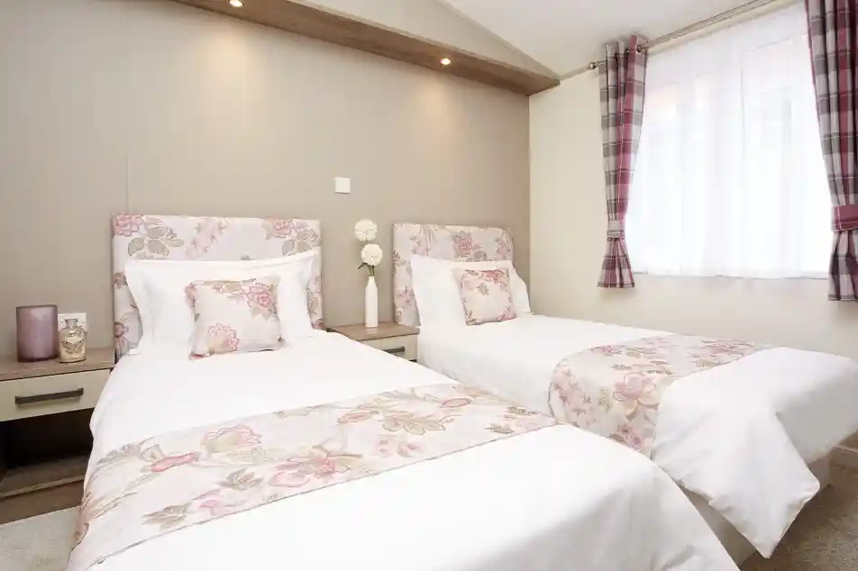 A cozy bedroom featuring two twin beds dressed in white linens with floral patterns. Soft pillows adorn the beds, and a warm-toned wall serves as the backdrop. Natural light filters through a window with sheer curtains, enhancing the inviting atmosphere.
