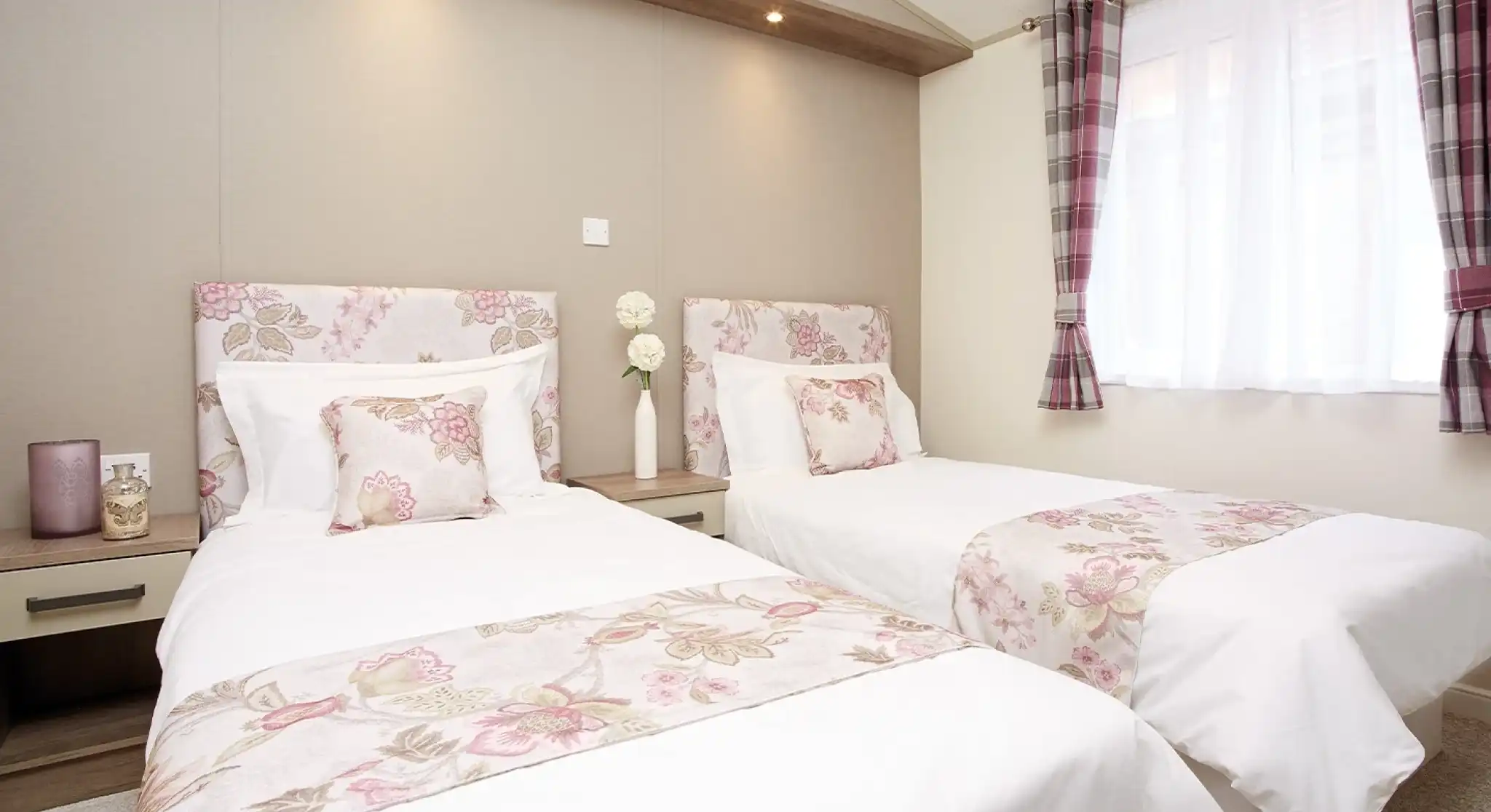 A cozy bedroom featuring two twin beds dressed in white linens with floral patterns. Soft pillows adorn the beds, and a warm-toned wall serves as the backdrop. Natural light filters through a window with sheer curtains, enhancing the inviting atmosphere.