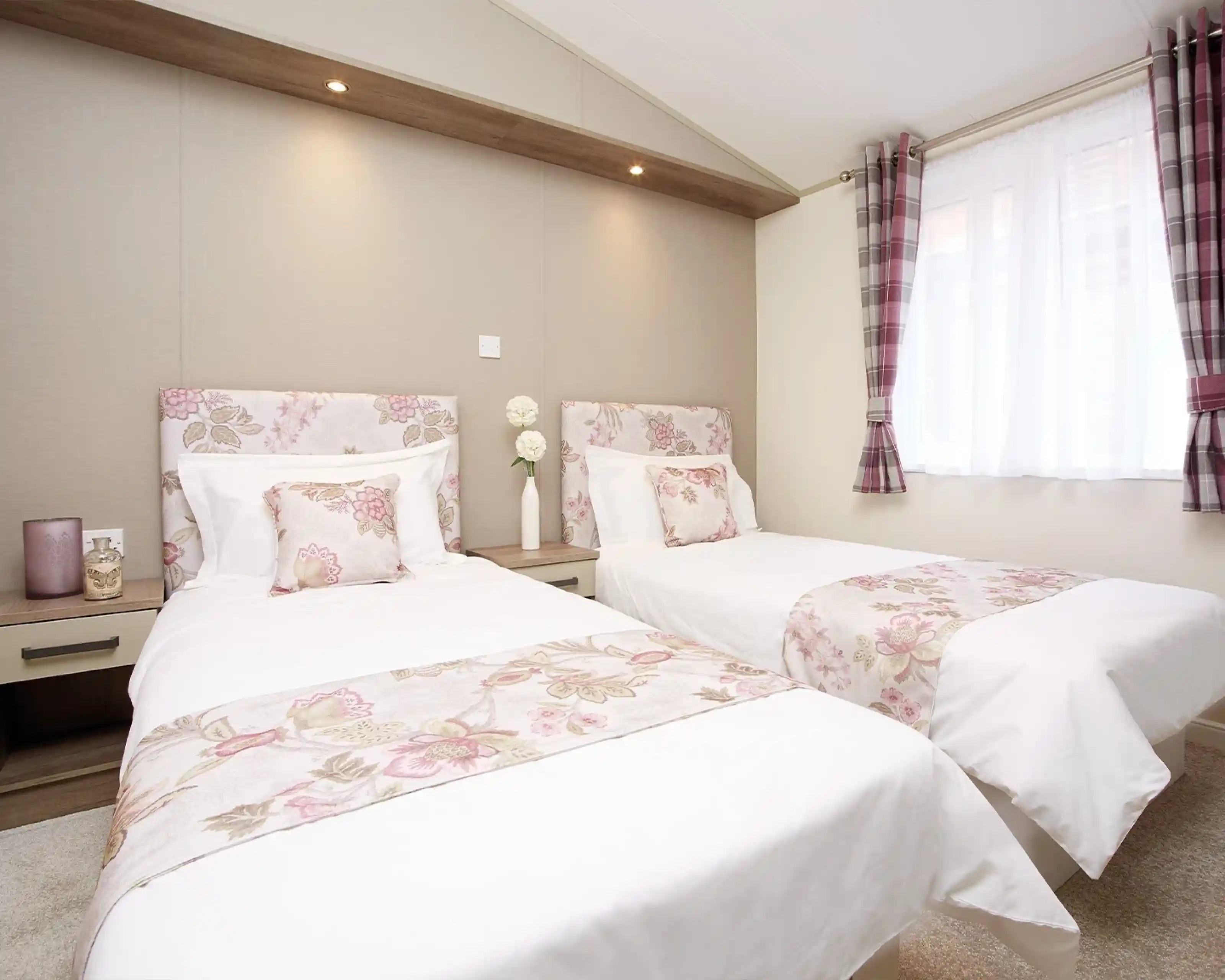 A cozy bedroom featuring two twin beds dressed in white linens with floral patterns. Soft pillows adorn the beds, and a warm-toned wall serves as the backdrop. Natural light filters through a window with sheer curtains, enhancing the inviting atmosphere.