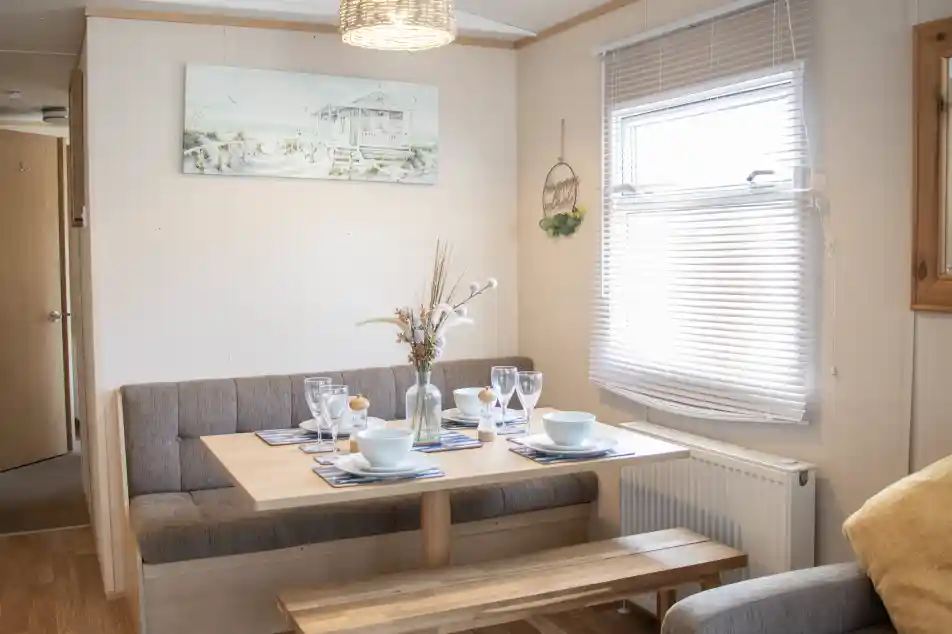 A cozy dining area features a wooden table set for a meal, with plates and glasses arranged. Soft seating is along one wall, and a decorative painting hangs above the table. Natural light streams through the window, highlighting a warm, inviting atmosphere.