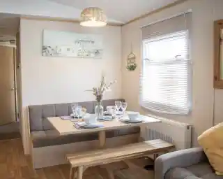 A cozy dining area features a wooden table set for a meal, with plates and glasses arranged. Soft seating is along one wall, and a decorative painting hangs above the table. Natural light streams through the window, highlighting a warm, inviting atmosphere.
