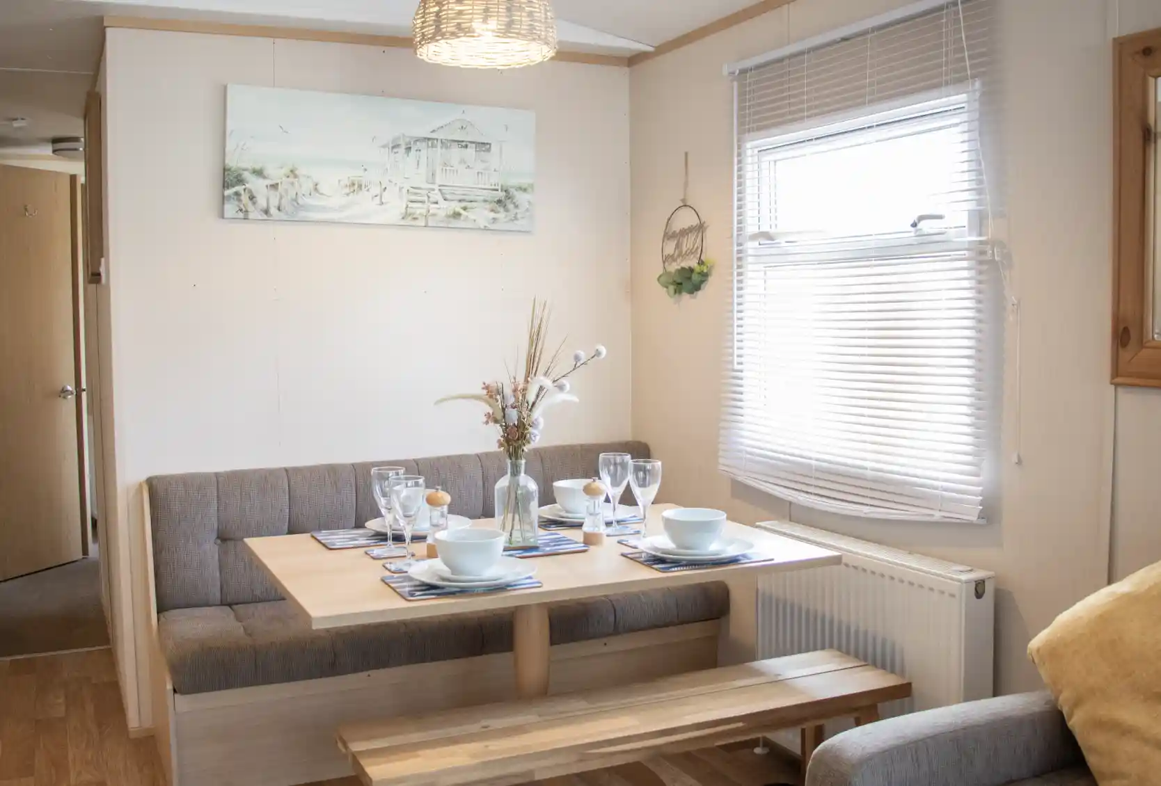 A cozy dining area features a wooden table set for a meal, with plates and glasses arranged. Soft seating is along one wall, and a decorative painting hangs above the table. Natural light streams through the window, highlighting a warm, inviting atmosphere.