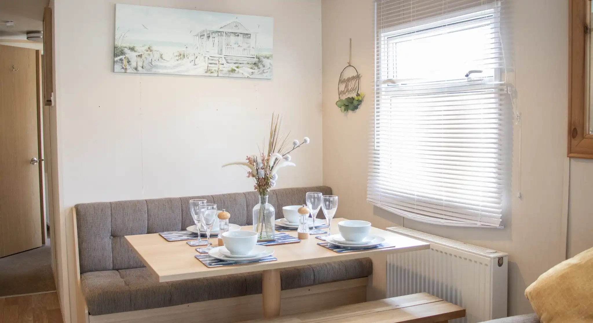 A cozy dining area features a wooden table set for a meal, with plates and glasses arranged. Soft seating is along one wall, and a decorative painting hangs above the table. Natural light streams through the window, highlighting a warm, inviting atmosphere.