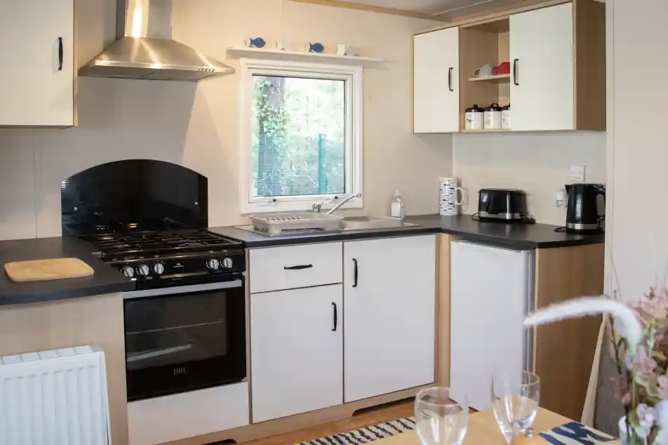 A modern kitchen featuring a black oven and stovetop, white cabinets, and a window overlooking greenery. Countertops are black, with a sink and coffee maker visible. A dining table is set with two wine glasses in the foreground.