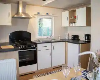 A modern kitchen featuring a black oven and stovetop, white cabinets, and a window overlooking greenery. Countertops are black, with a sink and coffee maker visible. A dining table is set with two wine glasses in the foreground.