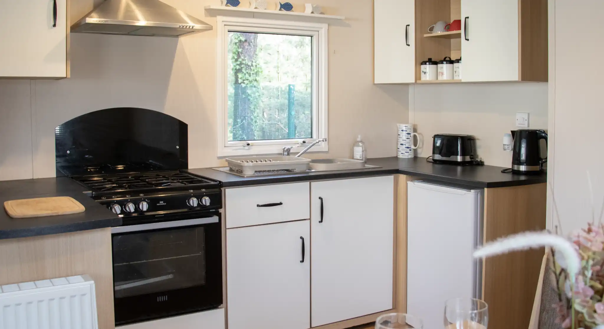 A modern kitchen featuring a black oven and stovetop, white cabinets, and a window overlooking greenery. Countertops are black, with a sink and coffee maker visible. A dining table is set with two wine glasses in the foreground.