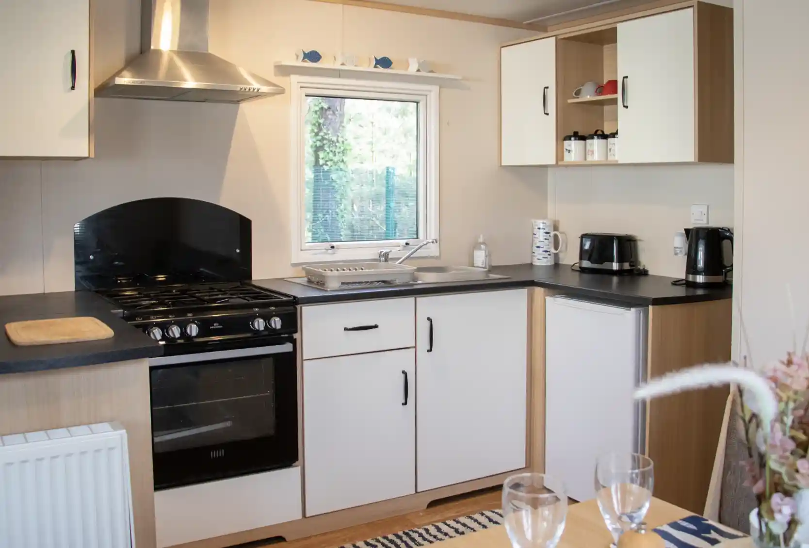 A modern kitchen featuring a black oven and stovetop, white cabinets, and a window overlooking greenery. Countertops are black, with a sink and coffee maker visible. A dining table is set with two wine glasses in the foreground.