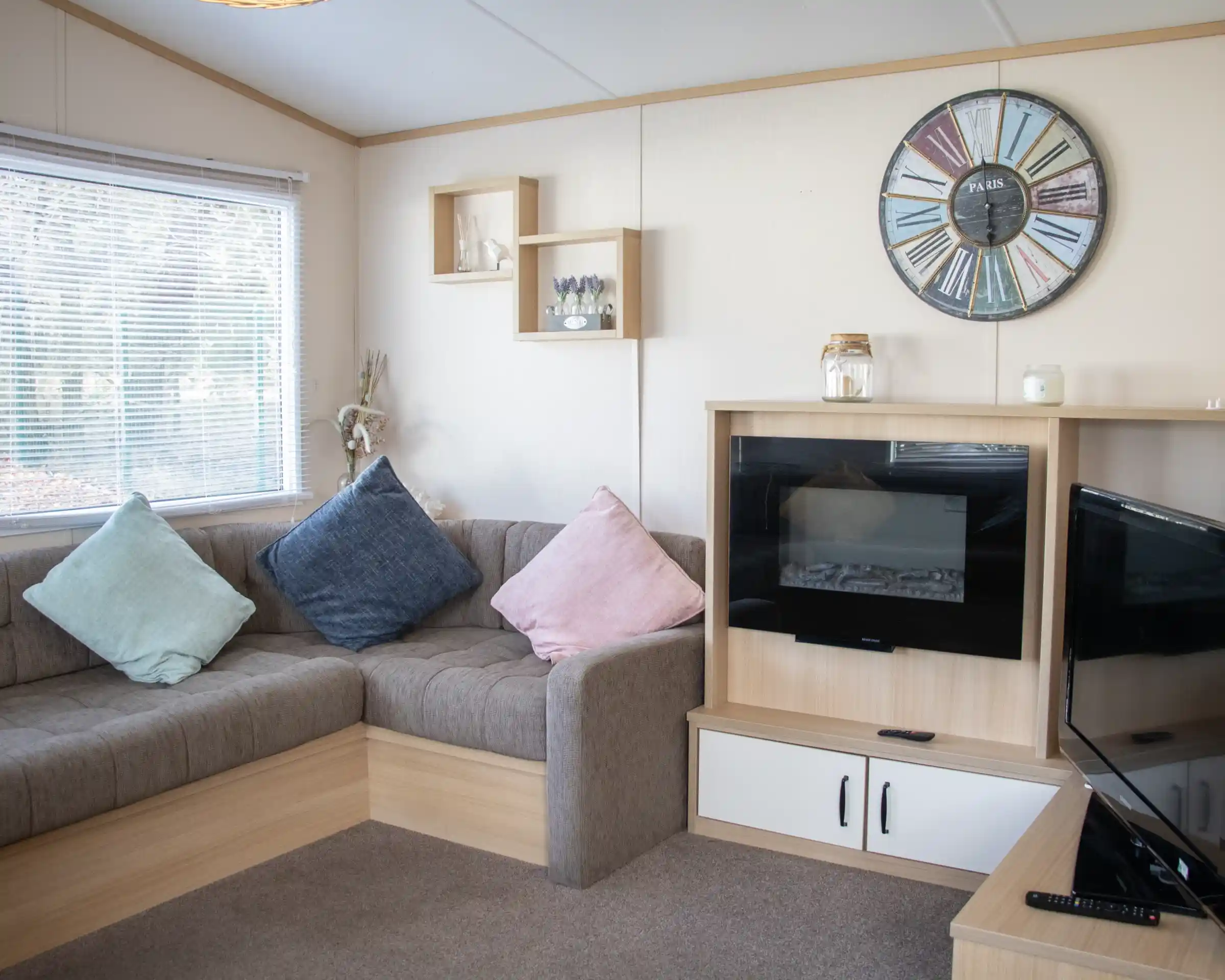 A cozy living room with a brown sectional sofa adorned with blue and pink cushions. A modern clock hangs on the wall, and there's a small shelf with decorative items. A fireplace is built into a wooden cabinet, next to a TV stand. Natural light filters through a window with blinds.
