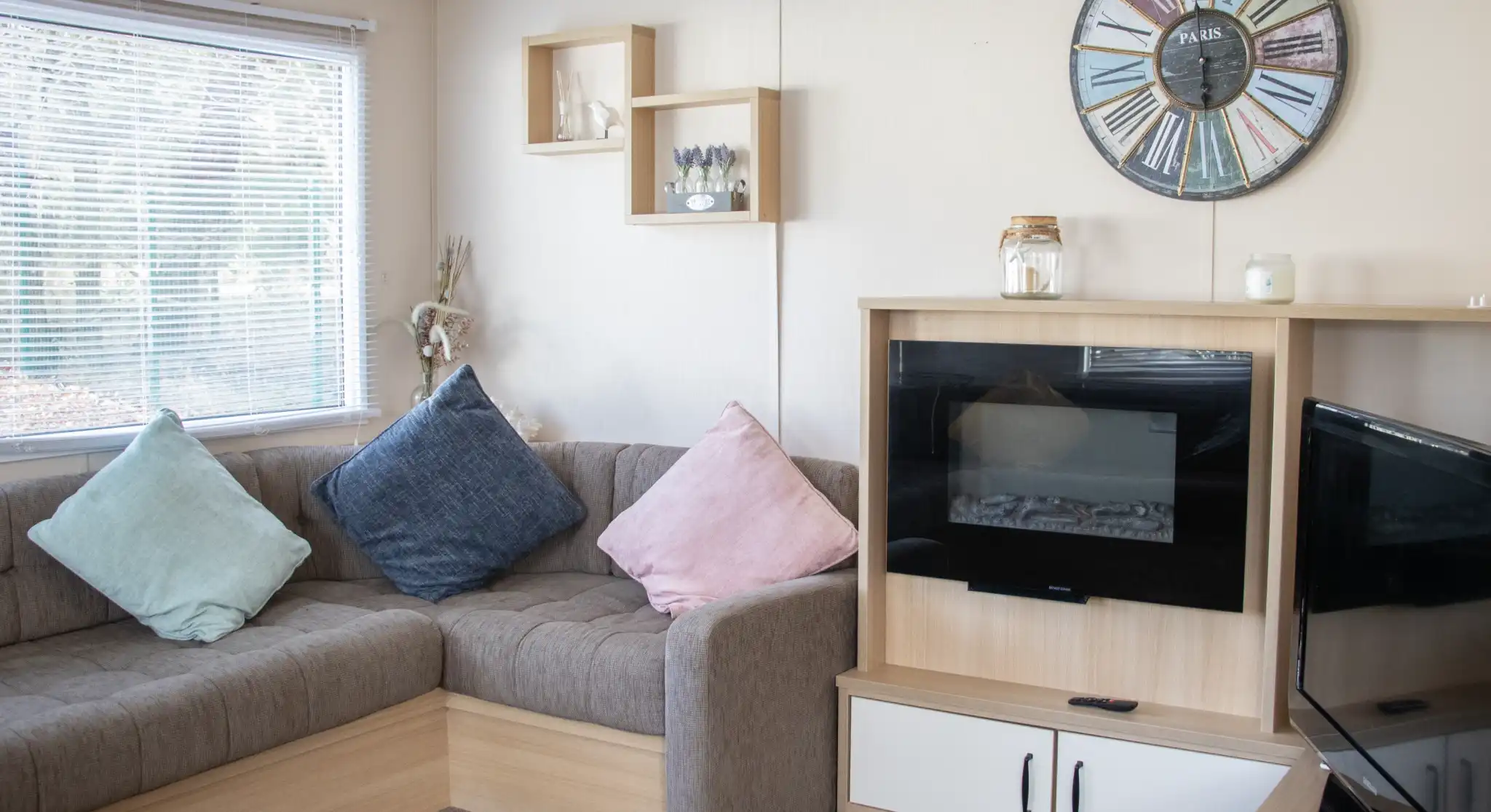A cozy living room with a brown sectional sofa adorned with blue and pink cushions. A modern clock hangs on the wall, and there's a small shelf with decorative items. A fireplace is built into a wooden cabinet, next to a TV stand. Natural light filters through a window with blinds.