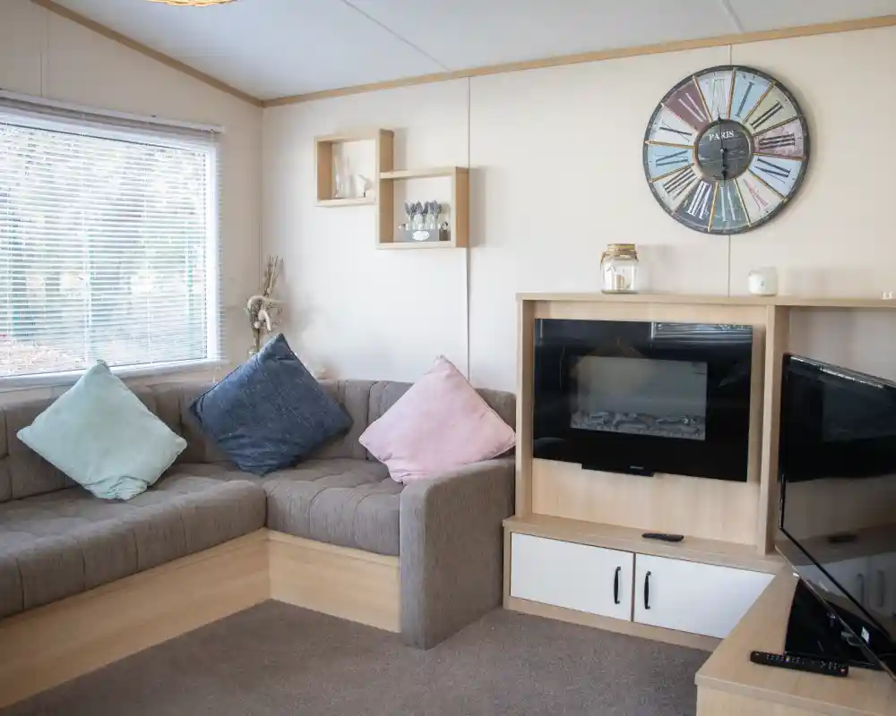 A cozy living room with a brown sectional sofa adorned with blue and pink cushions. A modern clock hangs on the wall, and there's a small shelf with decorative items. A fireplace is built into a wooden cabinet, next to a TV stand. Natural light filters through a window with blinds.
