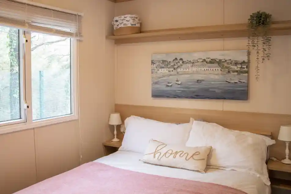 A cosy bedroom featuring a double bed with white linens and a pink throw blanket. A decorative pillow with the word "home" rests on the bed. There is a wooden shelf above the bed displaying a small plant and a decorative box. A large window offers natural light and greenery outside.