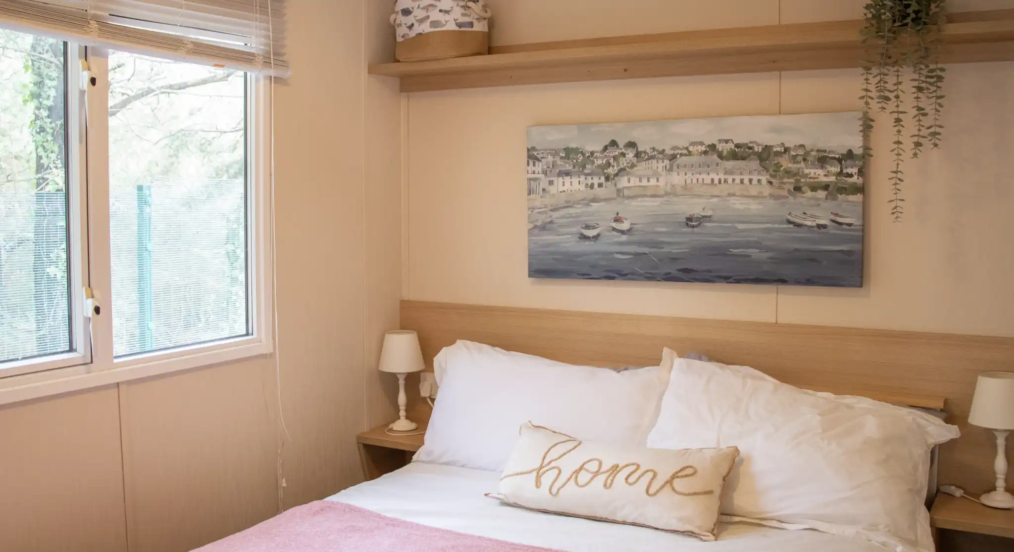 A cosy bedroom featuring a double bed with white linens and a pink throw blanket. A decorative pillow with the word "home" rests on the bed. There is a wooden shelf above the bed displaying a small plant and a decorative box. A large window offers natural light and greenery outside.