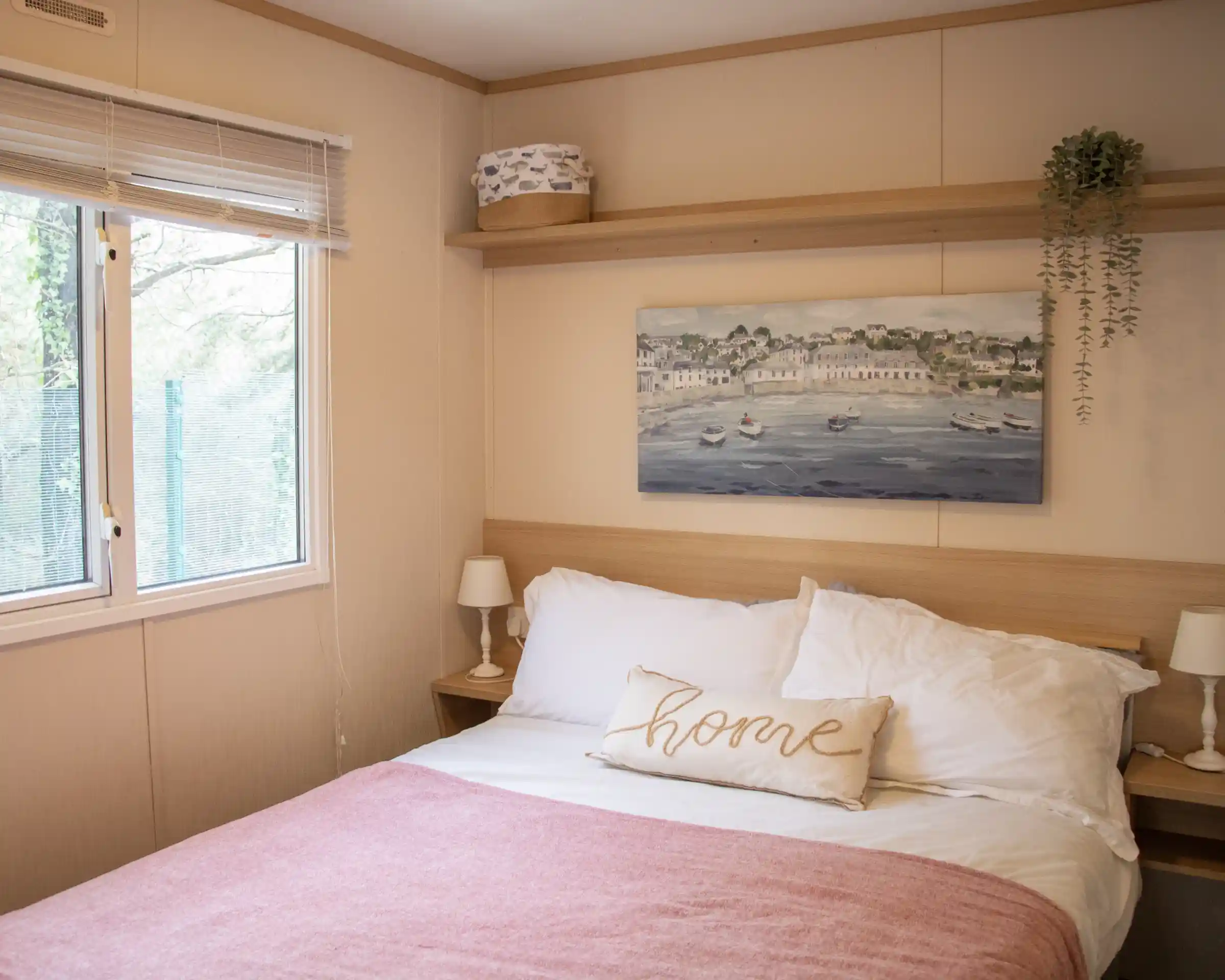 A cosy bedroom featuring a double bed with white linens and a pink throw blanket. A decorative pillow with the word "home" rests on the bed. There is a wooden shelf above the bed displaying a small plant and a decorative box. A large window offers natural light and greenery outside.