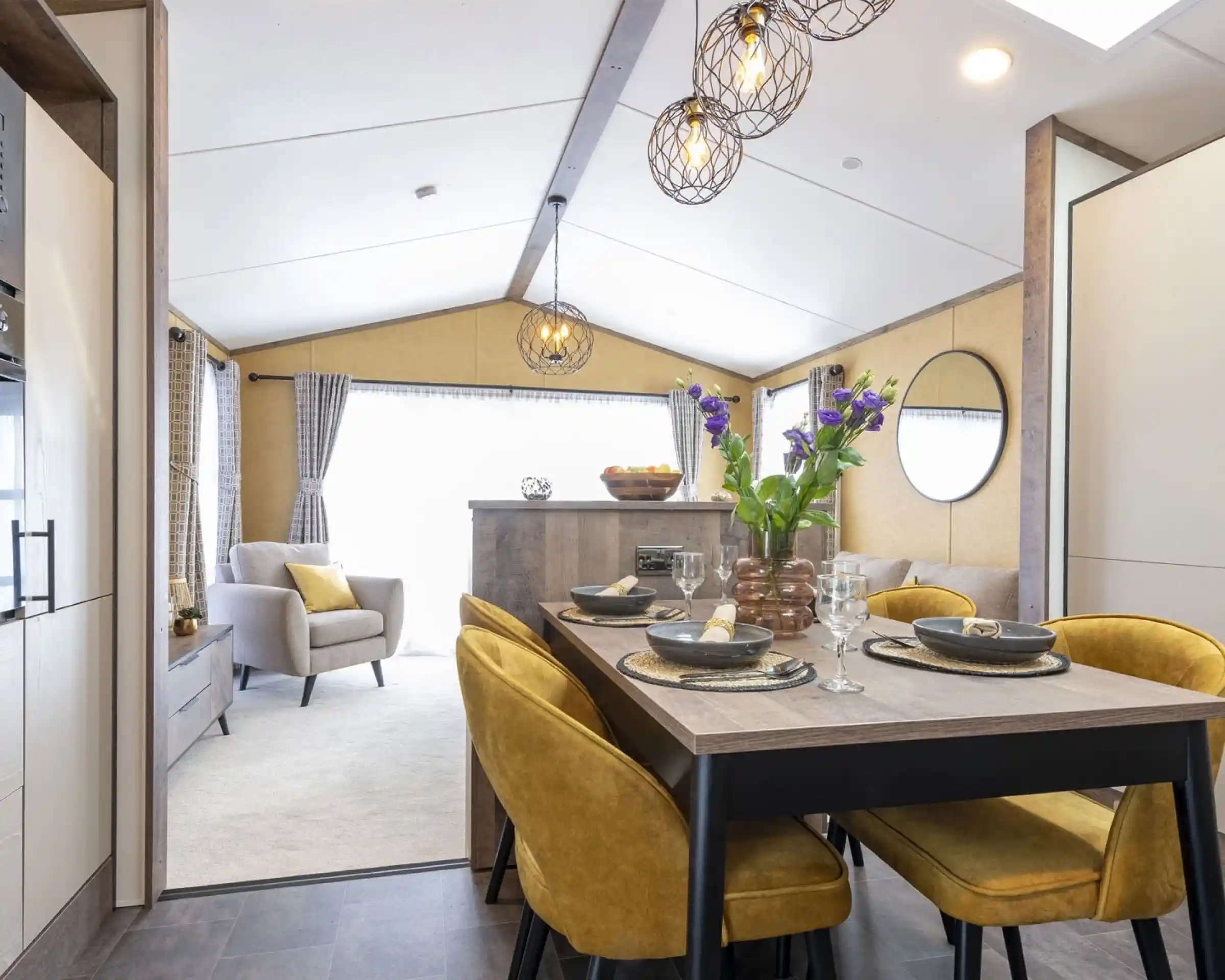 Cozy dining and living area with a wooden table, mustard-yellow chairs, and a vase of purple flowers. Light gray armchair and sofa in the background, with modern pendant lights and a round mirror on the wall. Bright and inviting space.